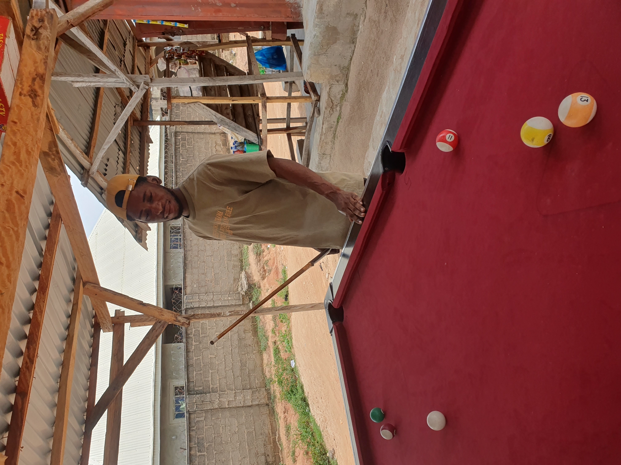 A young Black man wearing a tan t-shirt and a yellow baseball cap stands at a red felt pool table outdoors. He is holding a pool cue in his right hand, poised to strike a ball. Several pool balls are scattered across the table: a white cue ball, a solid green ball, a striped ball with the number 11, a striped yellow and white ball with the number 9, and a striped orange and white ball with the number 13. In the background, a rough wooden structure with a corrugated metal roof provides shade. Beyond the structure, a brick wall and a dirt path are visible, with some sparse vegetation. The scene appears to be in a casual outdoor setting, possibly a backyard or a local gathering spot in Lokoja, Nigeria.
