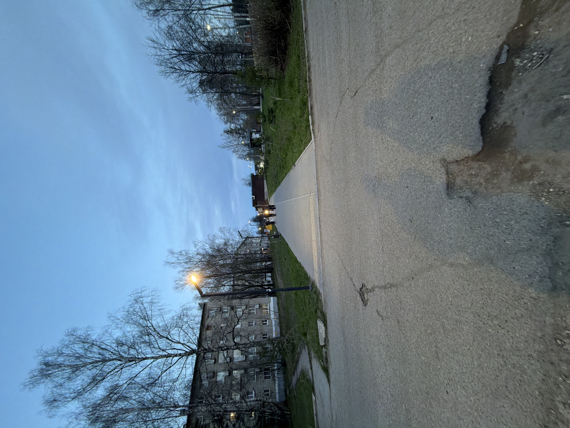 An eye-level, medium-range shot shows a paved sidewalk leading to apartment buildings in the city of Chaikovsky, Russia. The sidewalk is gray and cracked. To the right, the sidewalk is bordered by an eroded edge of asphalt. To the left, the sidewalk is bordered by grass. Beyond the grass are a series of gray apartment buildings. Bare trees surround the buildings. A street light illuminates the building on the left. A few people are walking in the distance. The sky is blue. The sun appears to be setting.