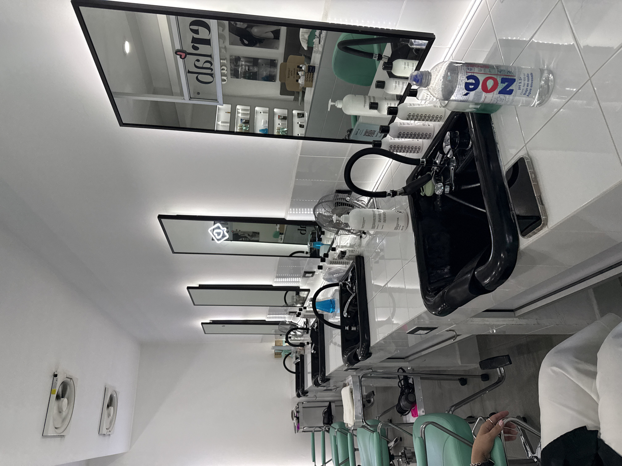The image captures an interior scene, likely within a salon or beauty parlor, in San Bernardino Tlaxcalancingo, Mexico. The focus is on a row of hair-washing stations, each equipped with a black basin, a flexible black hose, and a chrome faucet. Above each station is a rectangular mirror with a light bordering the edge. A mirrored wall reflects the salon interior. Several bottles of hair care products are visible on the counter of the first station. A clear water bottle with a blue cap and a label with the word "ZOE" is on the first counter. A smartphone is also next to the hair-washing basin on the first counter.
The salon has white walls and tiled counters. Ventilation fans are mounted on the wall, and mint green salon chairs are in front of each station. A person's arm with visible rings on the fingers is visible in the lower right corner of the frame.