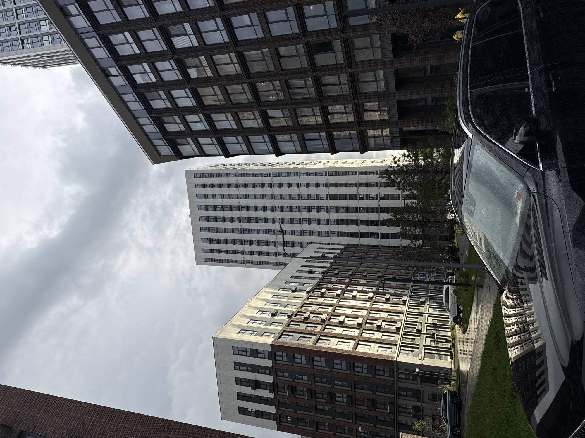 The photograph depicts an urban scene, likely in Moscow, Russia, as suggested by the architecture. The buildings are modern, high-rise structures with repetitive window patterns. One building is predominantly white with dark window frames, while others have a combination of dark brick and white panels. The sky is overcast with grey clouds. A black car is prominently positioned on the right side of the frame, with reflections visible on its wet surface. The composition is shot from a low angle, looking upwards towards the buildings and sky. The presence of foliage suggests a park or landscaped area adjacent to the road. Other vehicles are visible parked along the street, adding to the urban setting.