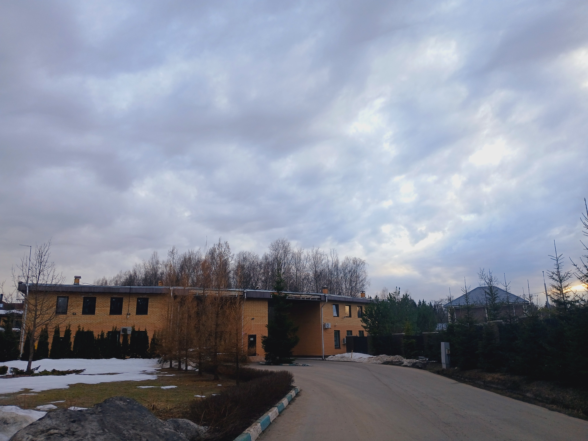 The scene depicts an outdoor environment under an overcast sky with patches of melting snow, suggesting late winter or early spring conditions. A two-story building, constructed of light brown brick with a dark, flat roof and several dark-paned windows, occupies the left-center of the image. Two chimneys are visible on its roof. An air conditioning unit is mounted on the exterior wall of the building's right section. Dark green evergreen shrubs are planted along the base of the building, particularly on its left side. Bare deciduous trees are situated behind the building and in various other spots.
In the foreground, an unpaved or asphalt road curves from the lower right towards the left, passing by the main building. The left edge of this road features a curb painted with alternating light blue and white sections. Patches of snow are visible on the brown grass and dirt areas adjacent to the road. A large, dark rock is present in the immediate lower-left foreground.
To the right of the road, a line of dense evergreen trees partially obscures a residential-style building with a dark, pitched roof. A utility pole or box stands near the road's edge on the right. The horizon on the far right shows a faint yellowish-orange glow, consistent with low sun visibility through the clouds. No individuals or active interactions are observed. The location is an unspecified outdoor area in Unknown, Unknown.