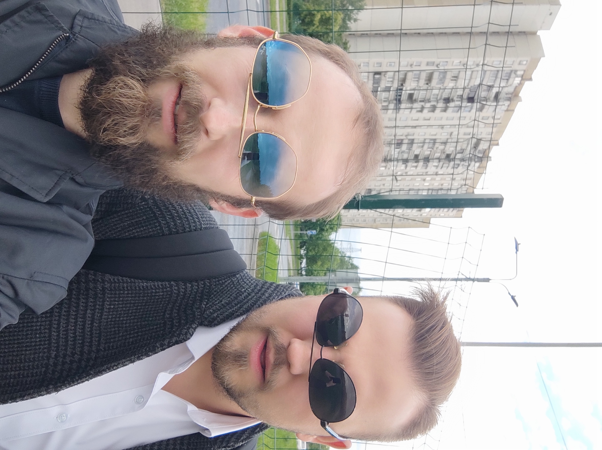 Two men take a selfie in front of a chain link fence. One man has a dark beard and wears gold-rimmed sunglasses. The other man is wearing a white collared shirt, dark blazer, and black sunglasses. Behind the fence, trees and a large apartment building are visible. This is in Moscow, Russia.