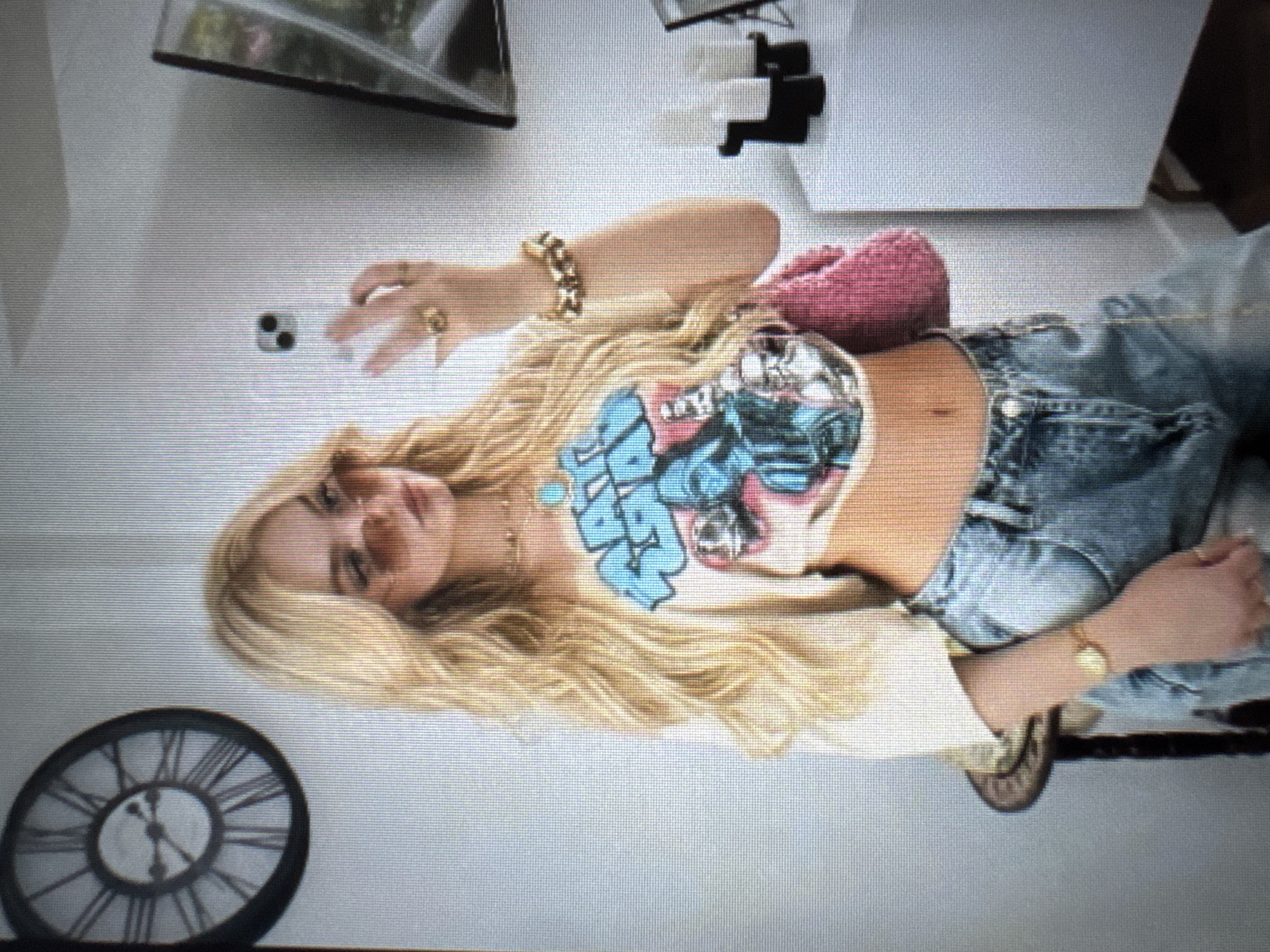 An image captures a young woman with long, flowing blonde hair, oriented diagonally across the frame. She is lying down, possibly on a bed or sofa, wearing a light-colored, cropped t-shirt with a prominent blue and white graphic design featuring stylized letters, possibly spelling "SWIM" or a similar word. Her midriff is visible. She is also wearing blue denim jeans.
The woman accessorizes with a gold chain necklace, multiple gold rings on her fingers, a thick gold bracelet on her left wrist, and a gold watch on her right wrist. A pair of sunglasses rests on her head, partially obscured by her hair. Near her right hip, a small, pink, knitted bag is visible. Her expression appears relaxed, and she is looking towards the viewer.
In the lower left corner of the frame, a round, dark-faced wall clock with prominent Roman numerals (e.g., X, XI, XII, I, II) is visible, with its hands indicating a time around 10:15 or 10:20. The background includes a portion of what appears to be a flat-screen television or monitor in the upper part of the image, and some white and black objects that resemble remote controls or decorative items. The entire image has noticeable horizontal scan lines, suggesting it is a photograph taken of a digital screen. The setting appears to be an indoor room, likely a living space or bedroom, under bright lighting.