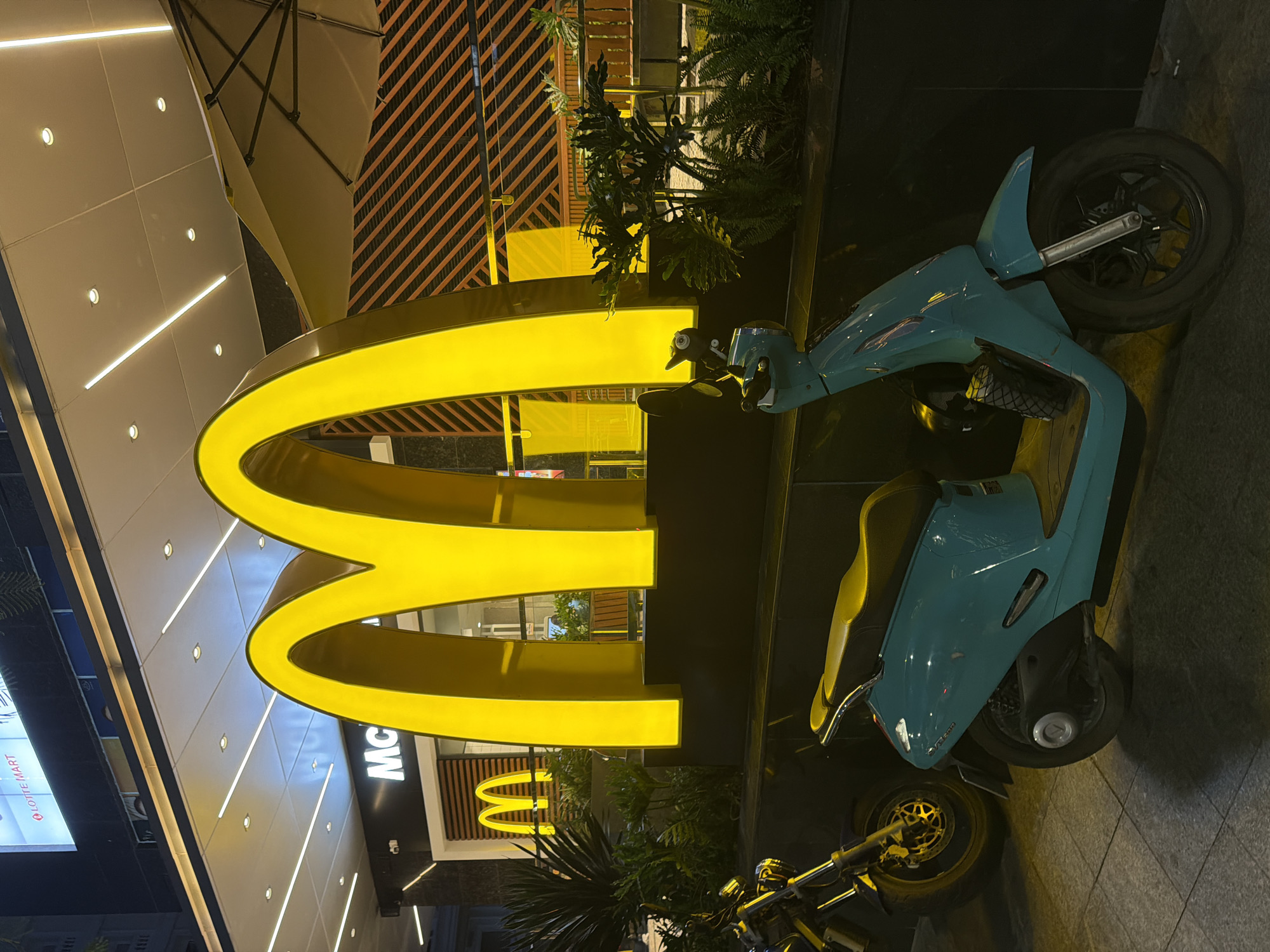 A bright, large yellow McDonald's arch logo illuminates the exterior of a McDonald's restaurant at night in Nha Trang Ward, Vietnam. Two scooters are parked in front of the restaurant. The scooter in the foreground is teal and faces towards the right, while a darker motorcycle is parked to its left. The restaurant's facade is lit with a modern, clean design, featuring white panels and recessed lighting. Behind the arch, orange diagonal stripes are visible on a wall, and a tan umbrella is partially visible. Lush green plants are strategically placed around the entrance. The ground is paved with grey tiles. The overall scene depicts a well-lit, modern fast-food establishment in a city setting during nighttime.