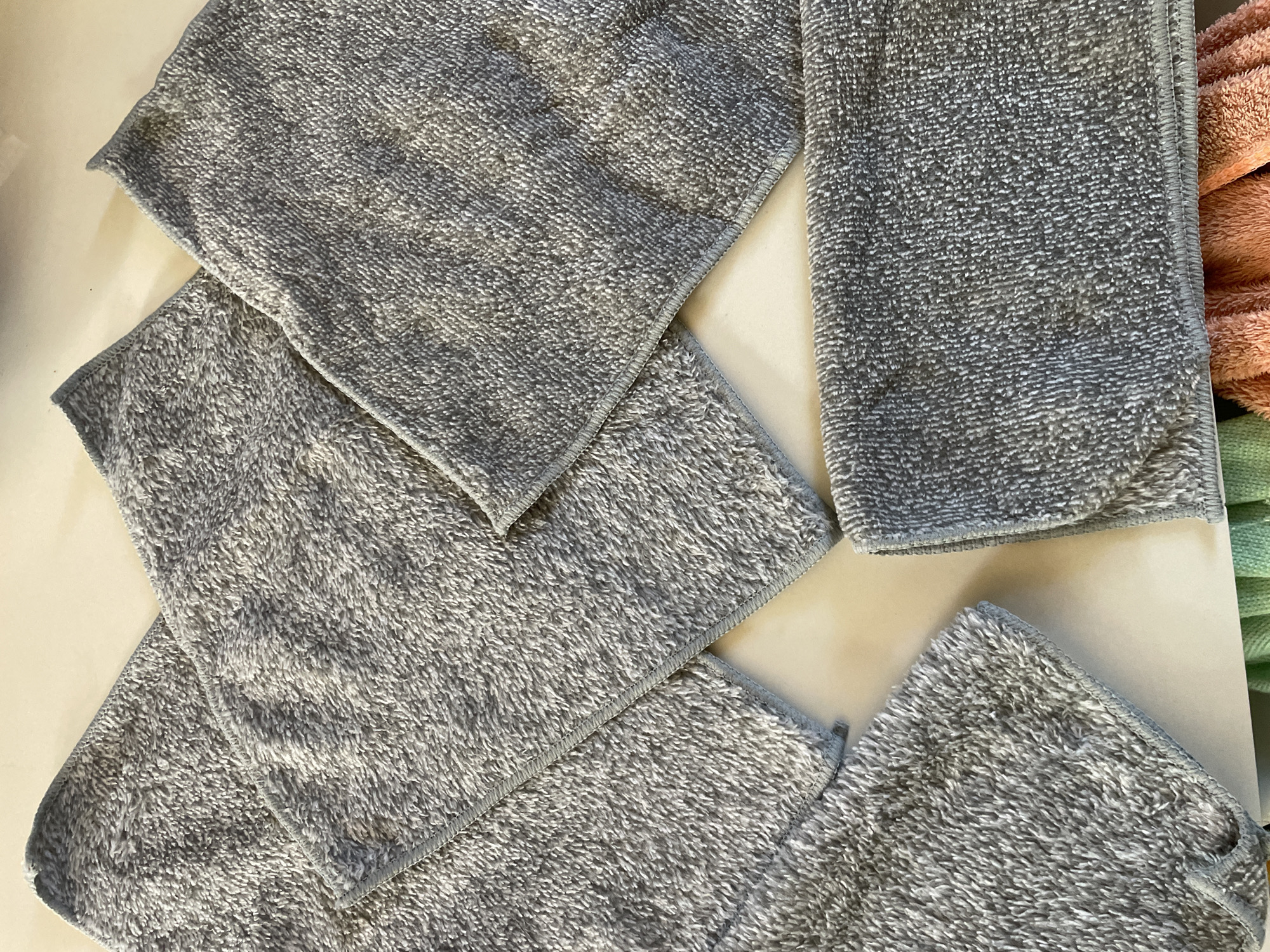 The image displays a close-up, overhead view of several grey microfiber cloths or towels arranged on a light-colored, possibly white or cream, flat surface. The cloths are unfolded and overlap each other in a somewhat scattered fashion, covering most of the frame. Their texture is visible, characteristic of microfiber, with a soft, slightly shaggy pile.
In the background, on the right side of the image, portions of other cloths or towels in different colors are visible and slightly out of focus. Specifically, an orange or peach-colored fabric is seen above a hint of green fabric.
No specific activity is taking place; the cloths appear to be laid out or piled. The lighting suggests an indoor setting. There are no people visible, and no text is present in the image. While the location context provided is Davao City, Philippines, there are no visual cues within the image itself to confirm this geographical location.
