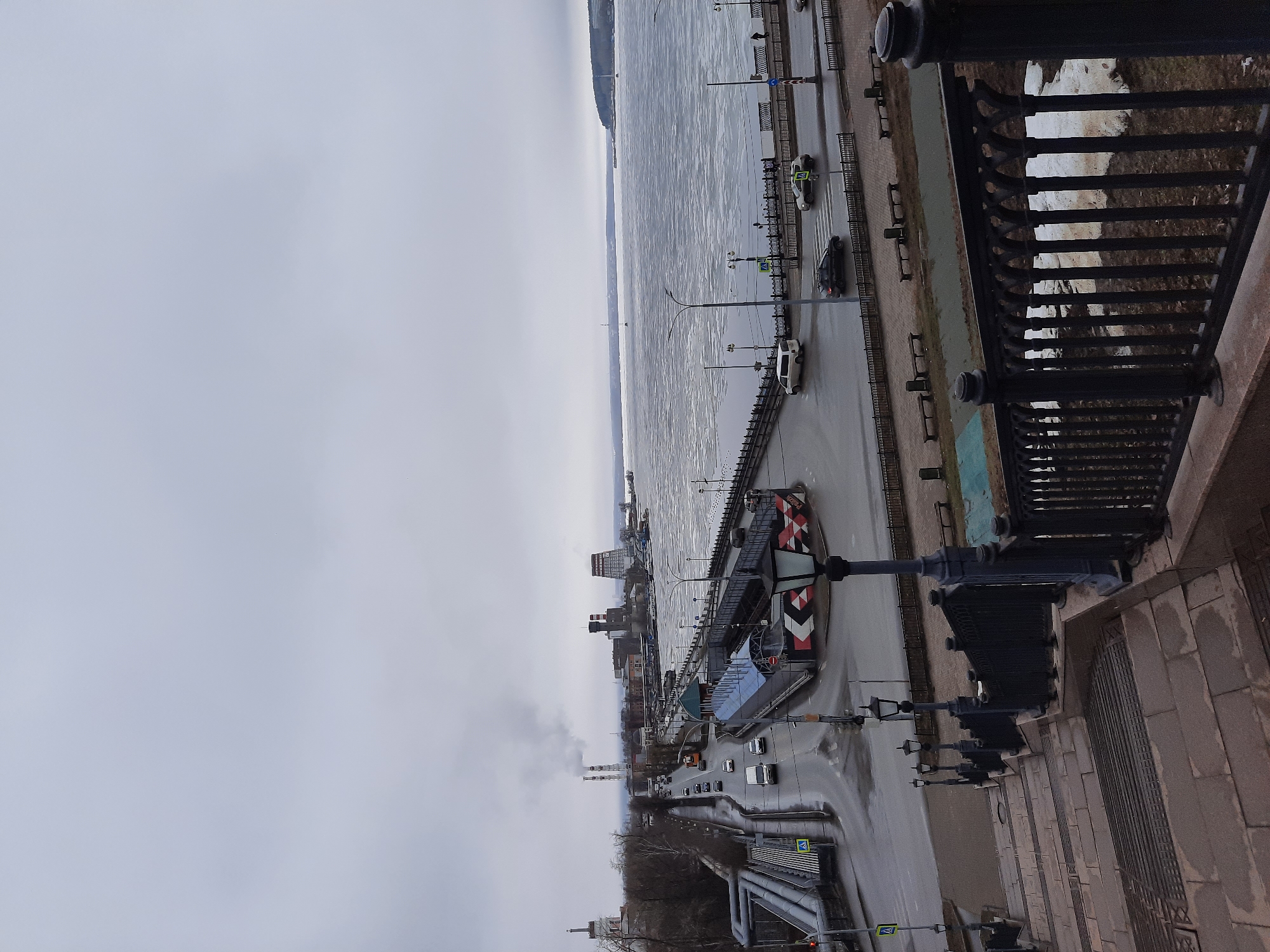 The image shows a view of the city of Izhevsk, Russia, from an elevated position, likely a bridge or high vantage point. A large body of water, possibly a river or lake, is partially covered with ice. Along the shoreline, there is a road with some vehicles, including cars and a truck, moving along it. There are also pedestrian walkways with benches. Buildings and industrial structures are visible further inland, with some smoke rising from chimneys, suggesting industrial activity. The sky is overcast, creating a muted color palette. The foreground shows a metal railing, indicating that the image was taken from a bridge or similar structure.