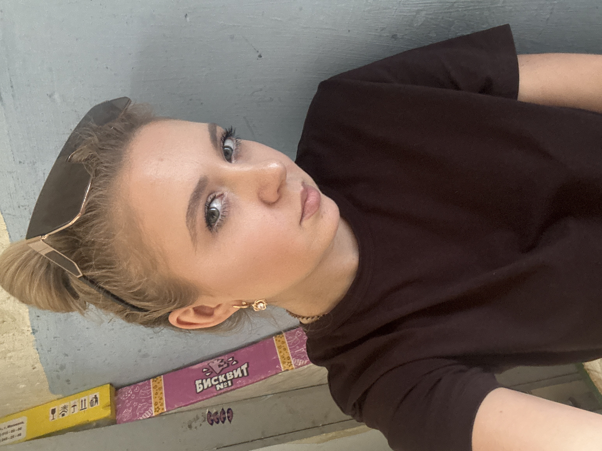 A young woman with blonde hair pulled into a bun and sunglasses perched on her head stands against a grey wall. She is wearing a dark brown t-shirt and a gold necklace. She has a gold earring with a small charm in her left ear. To her left, a pink and yellow box with Cyrillic lettering is leaning against the wall. The lighting suggests it is daytime.