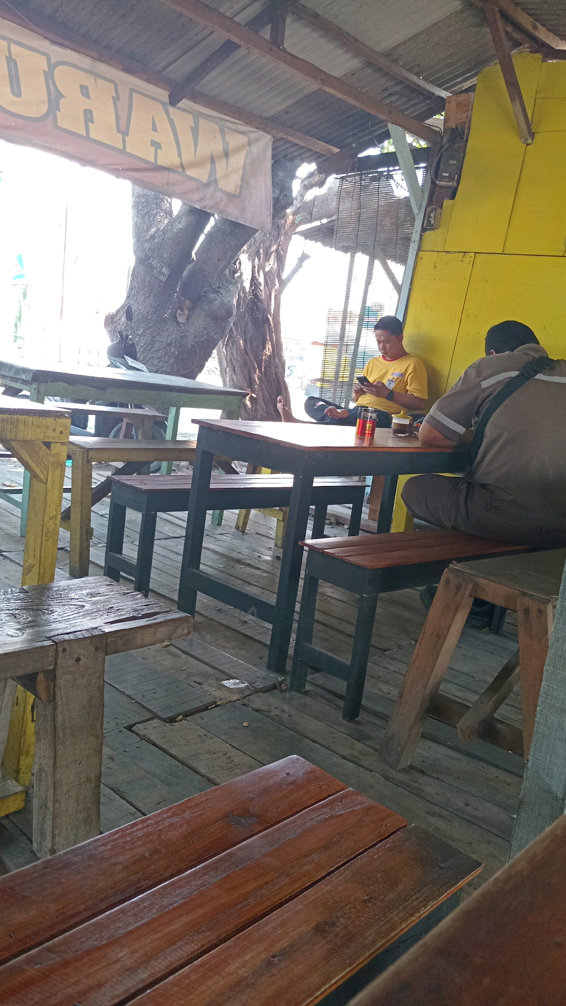 The image depicts an outdoor seating area at a warung in Ambeng Ambeng Watangrejo, Indonesia. Two men are present; one in a yellow shirt is seated at a table, looking at his phone. The other man, wearing a brown shirt with reflective stripes, is seated at the same table, facing away from the camera. On the table are drinks and possibly food. The warung appears to be constructed with a wooden floor and a corrugated metal roof supported by wooden beams. A large tree trunk is visible behind the table. The overall impression is a casual, rustic outdoor dining or resting spot.