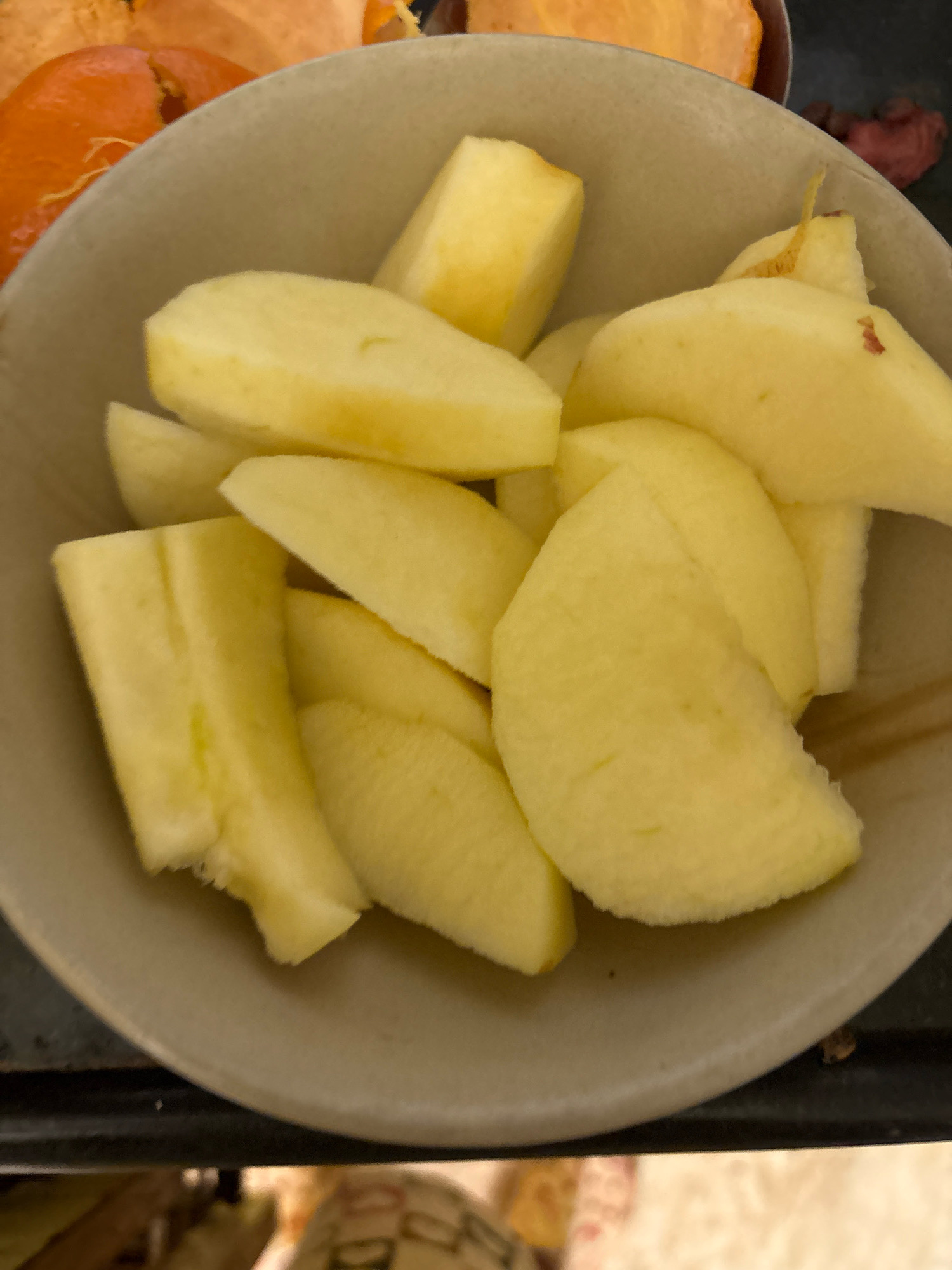 A close-up, overhead shot shows a bowl of sliced apples in Ashvem, India. The light-colored bowl is filled with apple slices, some of which appear to be slightly bruised or have small brown spots. Behind the bowl, out of focus, are slices of orange fruit, possibly persimmon or papaya, and a few dark red berries. The background is dark and blurry, making it difficult to discern any specific details of the environment.