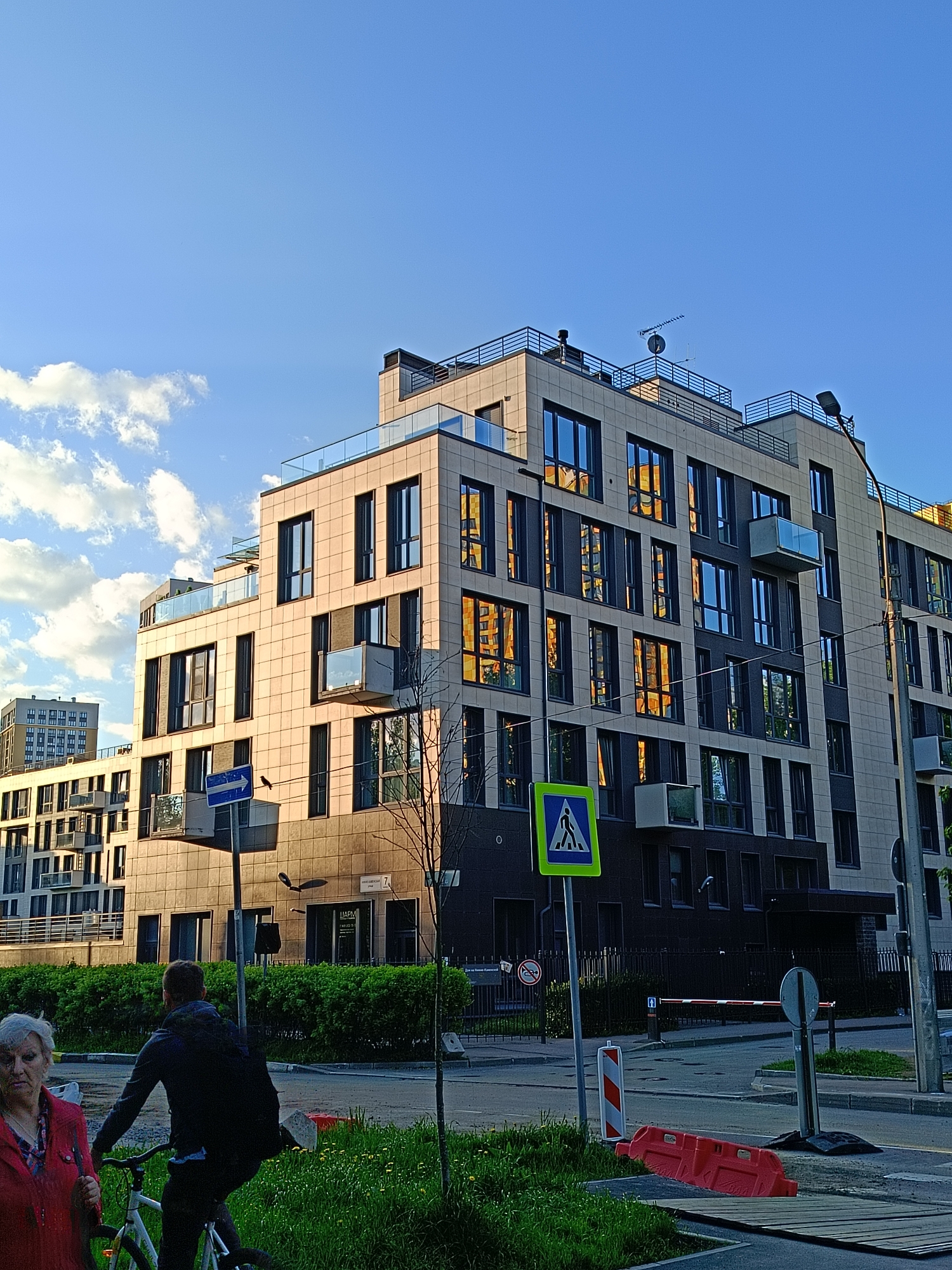 The image depicts a street scene in Saint Petersburg, Russia, featuring modern architecture and pedestrian activity. The main subject is a newly constructed multi-story residential building, characterized by a tile facade and large windows that reflect the golden light of the setting sun. The building features balconies with glass railings and rooftop installations including antennas and possible HVAC equipment.
In the foreground, a woman in a red jacket is walking to the left of the frame. A younger individual is on a bicycle. A pedestrian crossing sign and a street sign with an arrow are visible near the building. Further away, a gated entrance can be seen.
The street is lined with greenery, including bushes and grass. The sky is blue with scattered white clouds, indicative of fair weather. The scene is bathed in warm, late-afternoon sunlight. Another building is seen in the distance to the left. There are road barriers, including an orange barrier with white and red bollards.