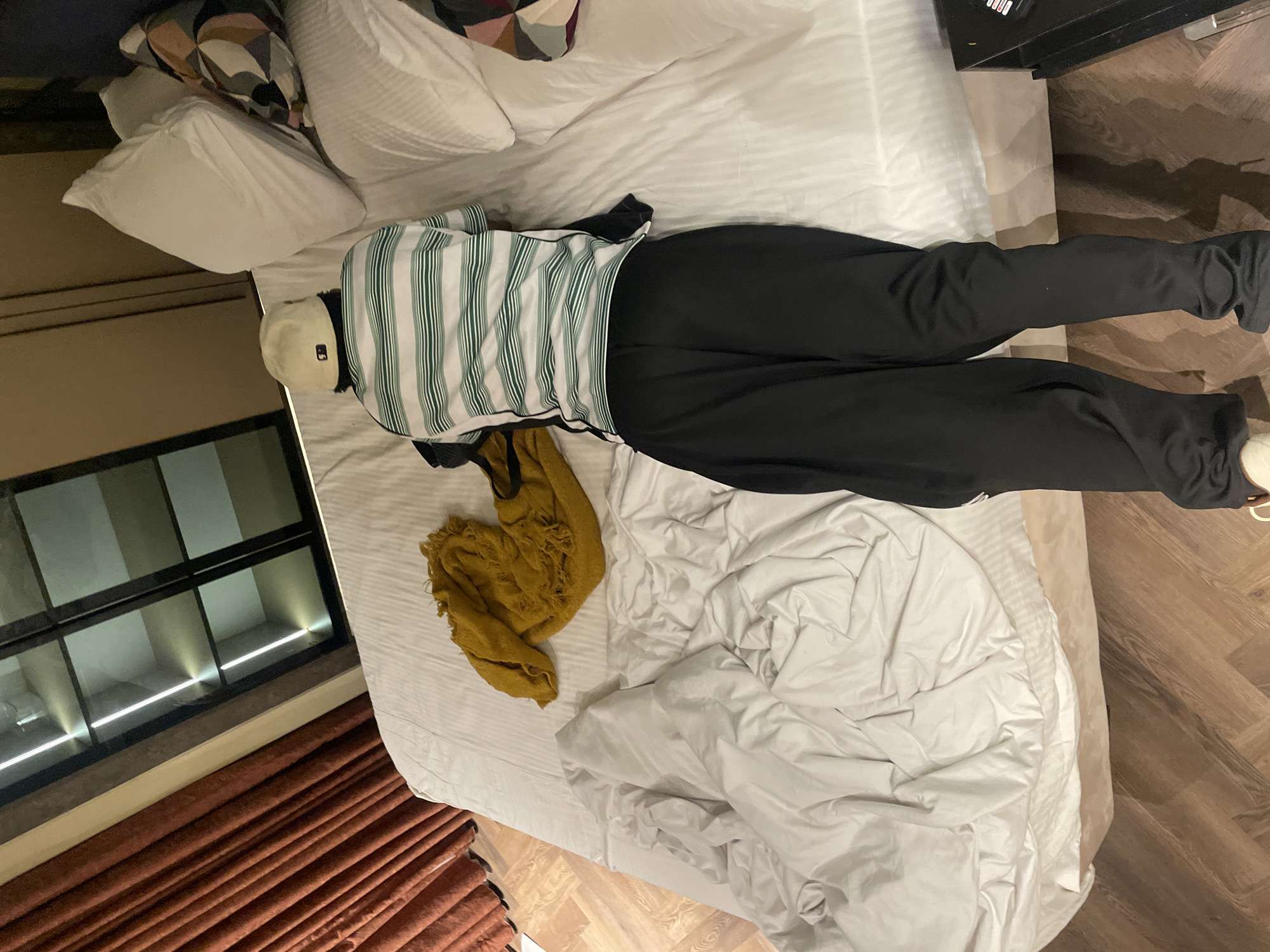 The image provides an overhead view of a person lying prone on a bed in what appears to be a hotel room or bedroom. The person is wearing a light-colored baseball cap, a white long-sleeve shirt with prominent green vertical stripes, and black pants. Their feet, clad in light-colored shoes, are visible at the bottom right of the frame.
The bed is covered with clean white sheets and features several pillows, one of which has a geometric pattern. A crumpled mustard-yellow garment, possibly a scarf or small blanket, is placed on the bed near the person's midsection.
To the left of the bed, a large, multi-paned window with a black frame reveals an architectural space beyond, illuminated by linear lights, suggesting evening or night. Below the window, brown pleated curtains are drawn. The floor on the right and bottom of the image is a wooden parquet with a herringbone pattern. A dark piece of furniture is partially visible at the top right.
The scene depicts a moment of rest or sleep. A small, dark rectangular logo is visible on the baseball cap, but no other explicit text is discernible. The overall ambiance is quiet and private.