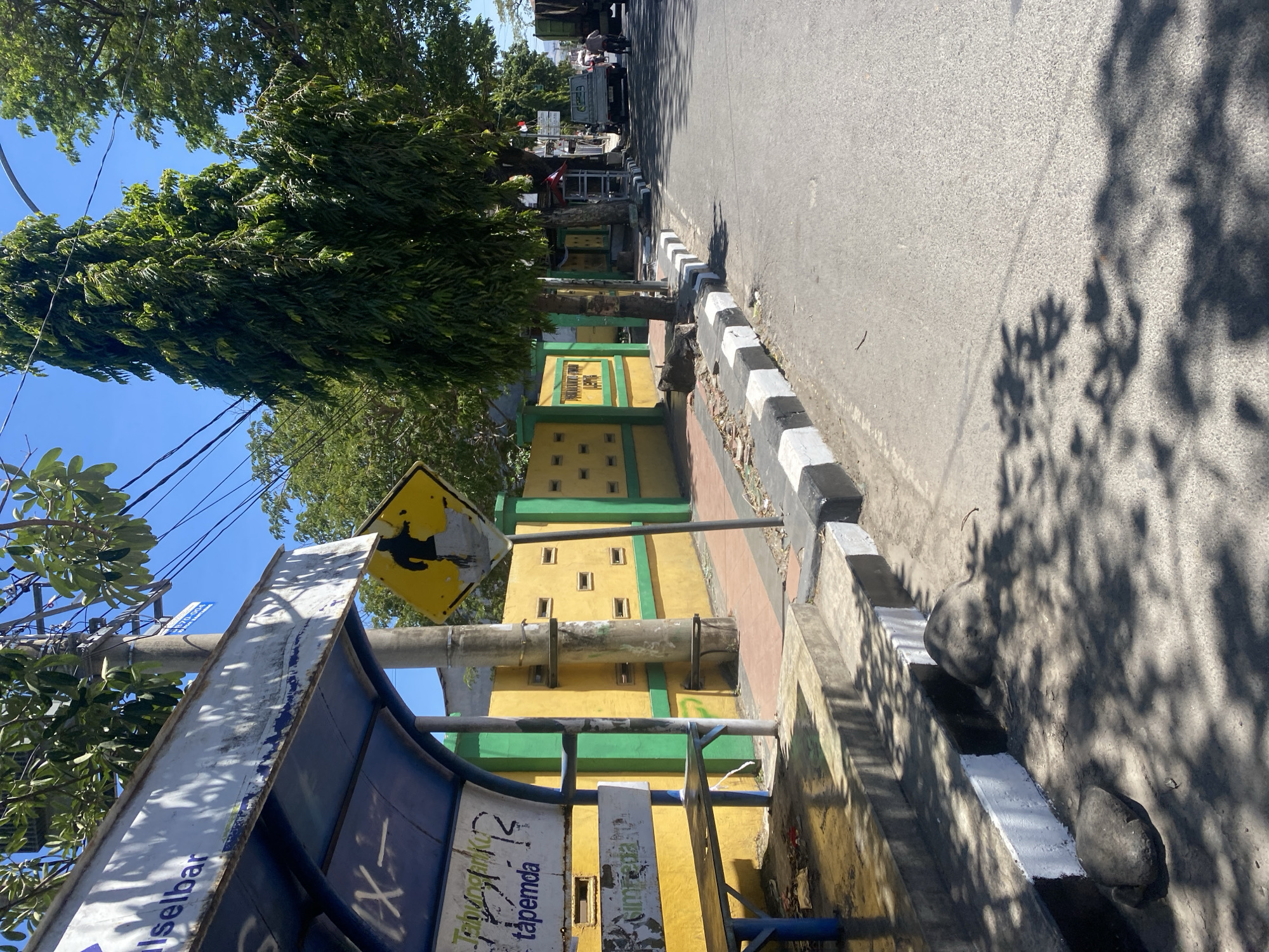 The image depicts a bus stop on a sunny day in Bantaeng, Indonesia. The bus stop features a blue roof with a weathered white sign on top, partially obscured by tree branches. A blue bench is visible inside the shelter, and the floor is concrete with debris. Adjacent to the bus stop is a bright yellow wall with green accents and a pattern of small rectangular openings. A pedestrian crossing sign with a silhouette of a person walking is posted on a pole near the bus stop. The street in front of the bus stop has a pavement with alternating black and white painted curbs. In the background, there are tall trees, power lines, and a street with some vehicles, including a truck and a motorcycle. The environment appears to be an urban or suburban setting in the city of Bantaeng, Indonesia.