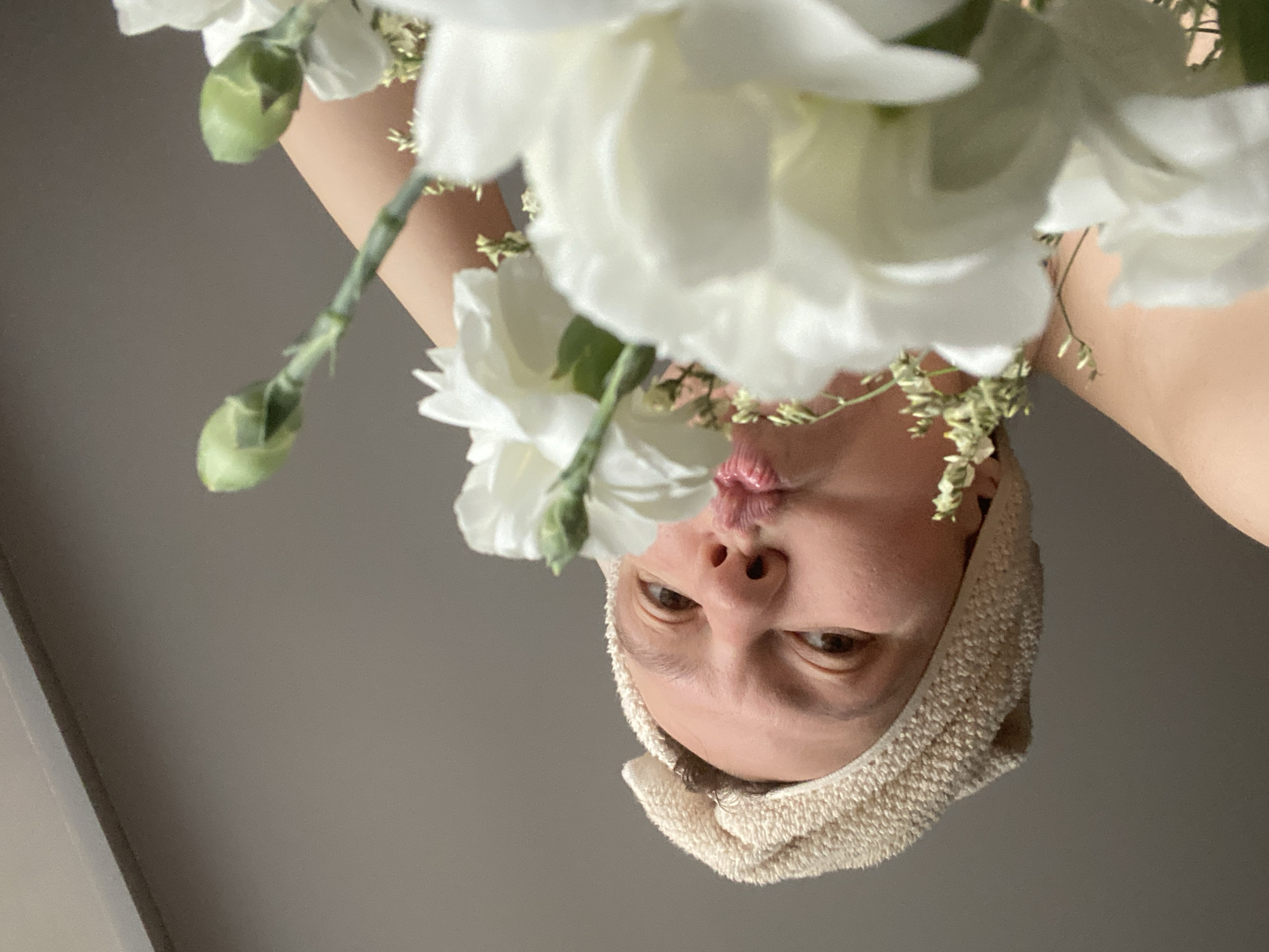 An indoor, close-up portrait shows an individual's face, positioned upside down, looking directly towards the viewer. The person has fair skin and is wearing a light beige or cream-colored towel wrapped around their head, resembling a turban or spa wrap. Their brown eyes are open and engaging, and their mouth is playfully pursed into a 'kissing' expression.
A bouquet of delicate white flowers, likely carnations, with slender green foliage, is prominently displayed in the upper half of the image, partially framing the person's face. Some flower blossoms are in sharp focus, while others are softly blurred. The flowers appear to be held near the person's face or just above them, creating an intimate composition. The upper part of the person's body, including a bare shoulder or neck, is visible in the upper right, suggesting a relaxed state.
The background is a plain, solid light grey or taupe surface, possibly a wall or ceiling, which minimizes distractions and keeps the focus on the subject. The lighting is soft and even, indicating an indoor setting with diffused light. The image conveys a whimsical and playful mood, capturing a moment of affectionate expression. This scene is set in Chaikovsky, Russia. No visible text is present.
