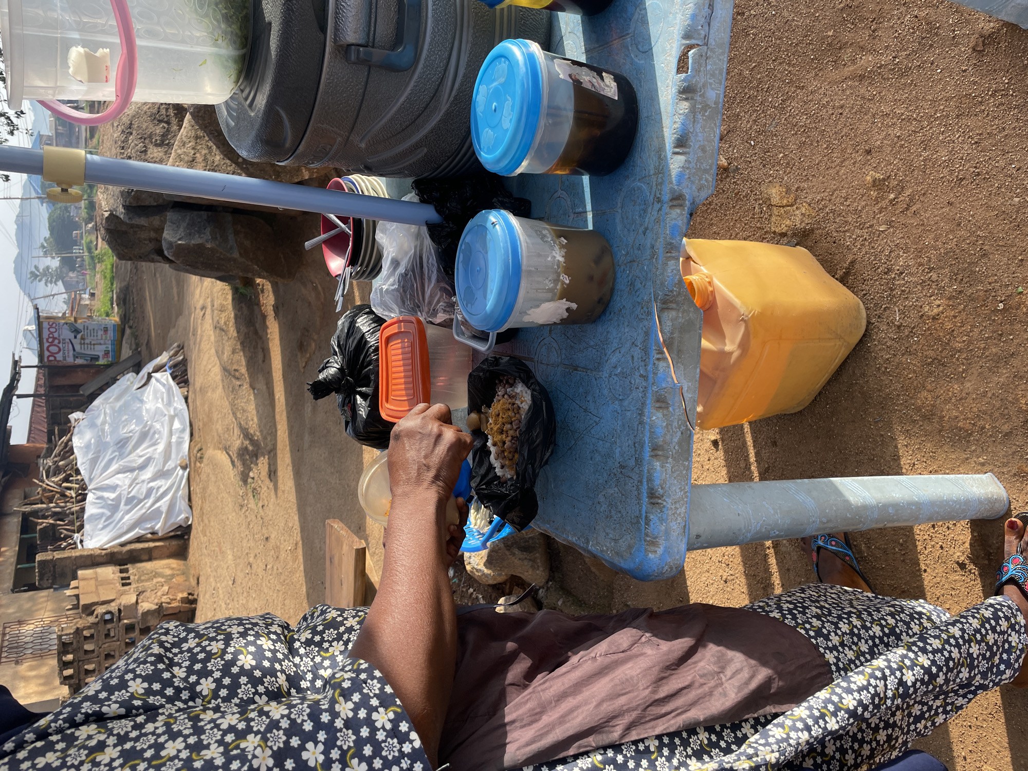 A woman wearing a patterned dress and brown apron is serving food from a table in an outdoor market. She is holding a plastic bag with food in it, and her hands are visible. On the table, there are various containers of food and drinks, including plastic cups with blue lids and a large jug. In the background, there are buildings, rocks, and a pile of wood. The ground is dirt and sand. The scene appears to be a food stall in a market in Jos, Nigeria.