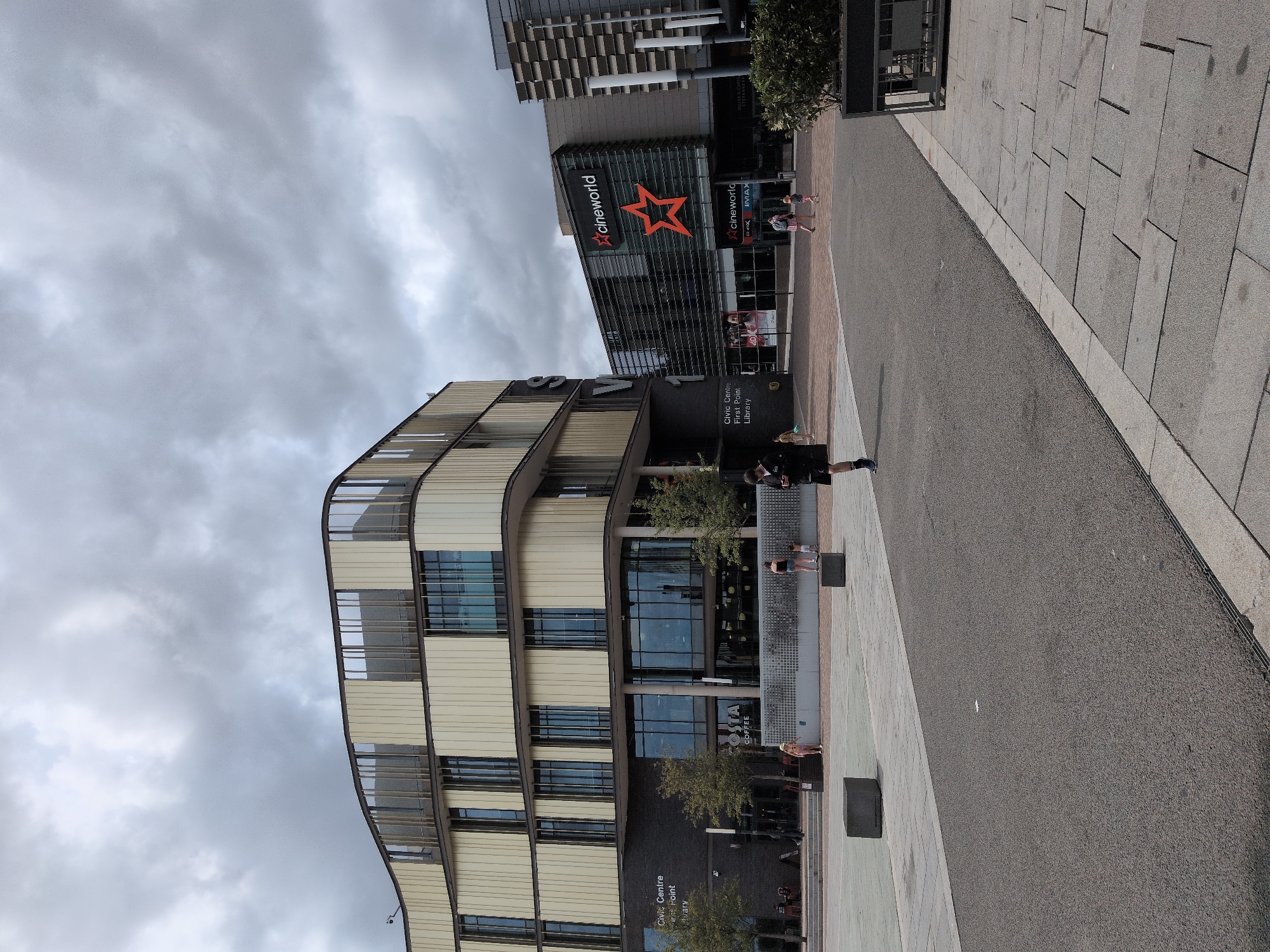 The image depicts an outdoor urban scene in Telford, United Kingdom, featuring modern architecture. On the left, a curved, multi-story building with beige facade panels and dark-framed windows dominates the foreground. The ground floor has large glass windows, with signage indicating "Civic Centre," "First Point," and "Library." Adjacent to this building, to the right, is a darker building with the "Cineworld" logo prominently displayed above its entrance, featuring a large orange star graphic. This Cineworld building has multiple levels of glass facade with visible internal structures and entrances.
In the central area, a paved plaza is visible with several large, dark grey concrete blocks placed around the space. A few individuals are present: a person in dark athletic wear stands on the right side of the plaza, facing away from the camera. Further into the plaza, two children, one in a pink outfit and another in a light-colored outfit, are walking. Behind them, an adult female in a black tank top and shorts is walking with a younger child.
Trees are sparsely planted in planters along the facade of the Civic Centre building. The sky is overcast with heavy grey clouds, suggesting a cloudy or potentially rainy day. The ground in the foreground is a dark asphalt surface with a lighter paved sidewalk to the right. The overall impression is a modern civic and entertainment complex in a town square.