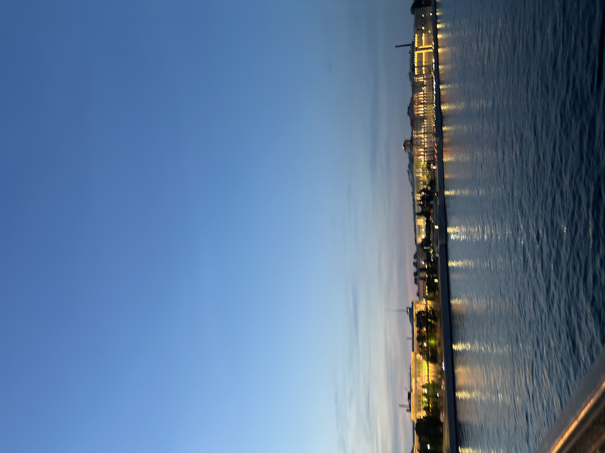 The image depicts a serene evening scene along the embankment of the Neva River in Saint Petersburg, Russia. The sky above is a deep, clear blue, transitioning to lighter hues near the horizon, indicating the time is dusk or early night. Wispy clouds are visible, adding subtle texture to the sky.
Along the far bank of the river, a long, grand building complex is illuminated with warm, yellow lights. The architecture suggests historical significance, with multiple windows and a prominent dome-like structure visible. The lights from the building are reflected in the dark, rippling waters of the Neva, creating a shimmering, elongated pattern on the surface.
Trees are planted along the embankment in front of the building, their dark silhouettes contrasting with the illuminated facade. A few indistinct shapes that could be vehicles are visible on a road running along the riverbank. The foreground shows the dark, rippling surface of the Neva River, with a hint of a railing in the lower left corner, suggesting the viewer is on a bridge or a viewing platform. The overall atmosphere is one of peacefulness and grandeur, characteristic of Saint Petersburg at twilight.