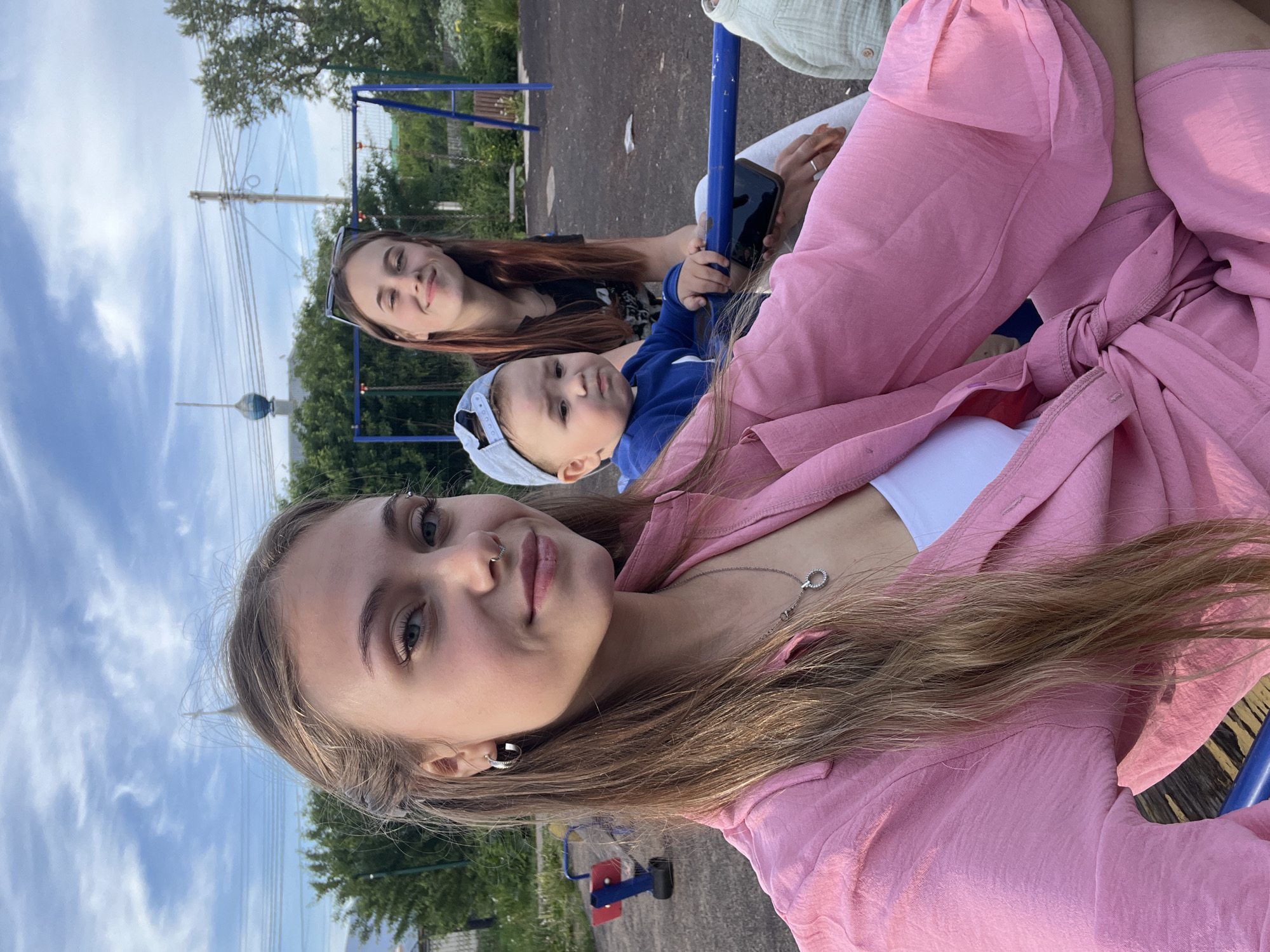 The image depicts three individuals – two young women and a baby – enjoying an outdoor setting, likely a park or playground in Фоки, Russia. The photo appears to be a selfie taken by the young woman in the foreground.
This woman has long, light brown hair, light eyes, and wears a nose piercing and earrings. She is dressed in a pink long-sleeved top or jacket over a white garment, accessorized with a silver necklace, and looks at the camera with a slight smile. Behind her, a second young woman with long, reddish-brown hair, wearing sunglasses pushed onto her head and a dark top, is also smiling and appears to be holding a smartphone. Between the two women sits a baby or toddler, wearing a light blue baseball cap backwards and a blue long-sleeved top, looking towards the camera with a neutral expression.
The group is positioned near a blue metal structure, resembling part of a swing set. The background features lush green trees and other playground equipment in red and blue. The sky is bright with scattered white clouds, indicating clear daylight. A utility pole with multiple power lines is visible, and in the distant left background, a distinctive spherical, dark-colored structure on a pole or building stands out, potentially a weather radar or similar landmark. The overall scene conveys a relaxed and pleasant atmosphere. No visible text is present in the image.