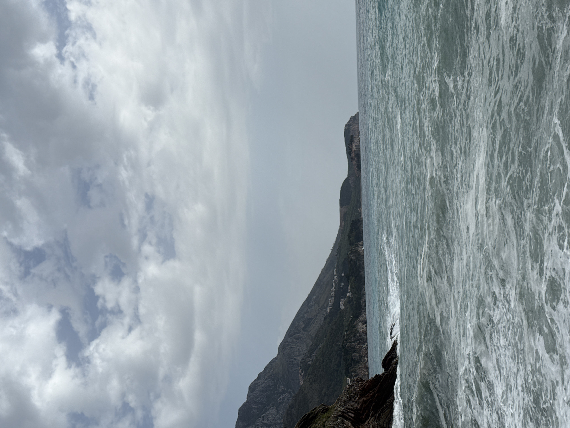 The image shows a mountain range meeting the ocean under a cloudy sky. The mountain is dark and rocky, with some greenery visible. The ocean is a turbulent grey, with white foam on the surface. The location is Unknown, Unknown.