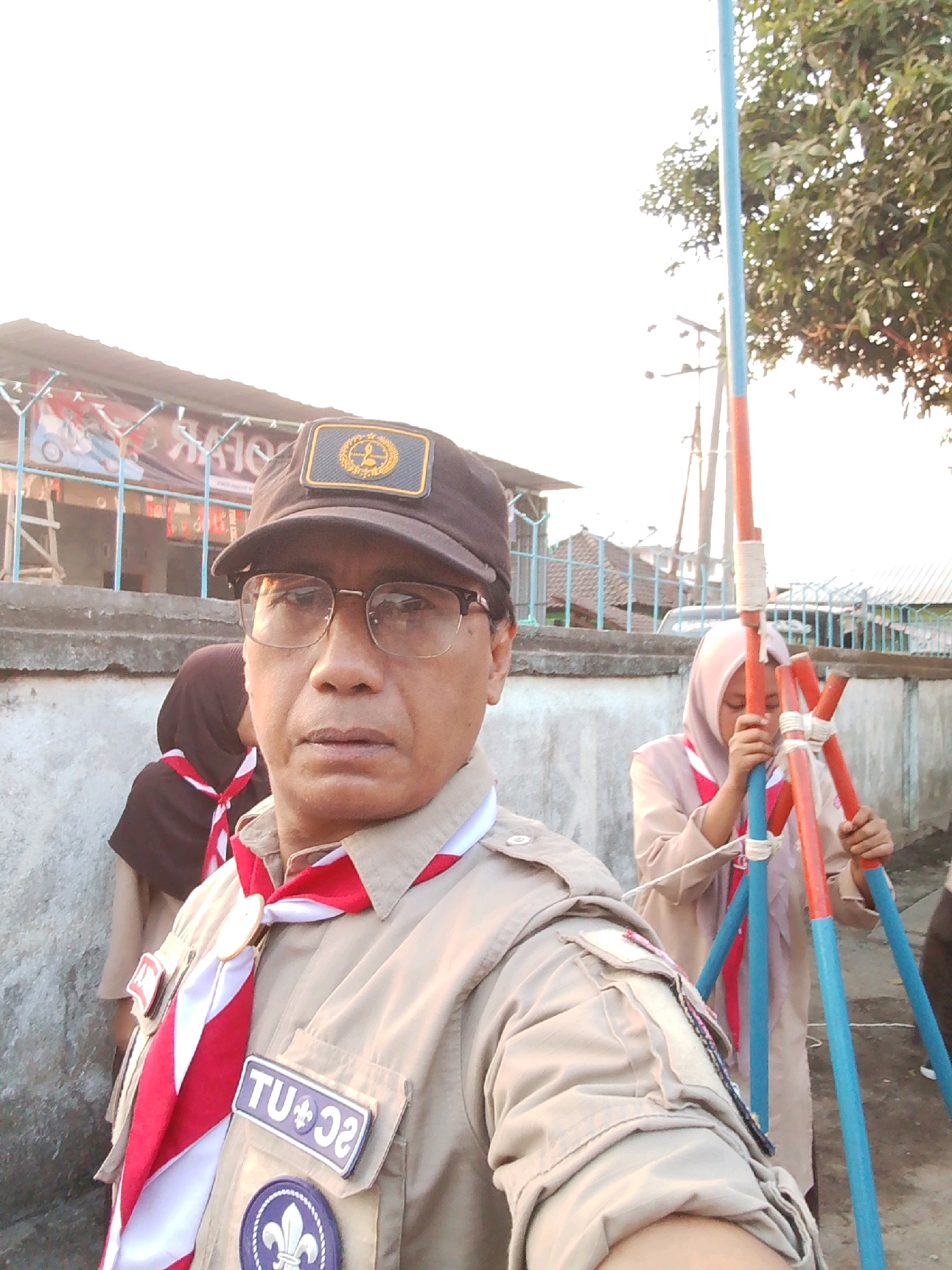 In Kuripan Utara, Indonesia, a man wearing a brown scout uniform, a cap, and glasses takes a selfie outdoors. He is standing in front of a building with a textured wall, and a fence is visible behind him. Two young girls in scout uniforms are also present. One girl, wearing a brown hijab and uniform, stands to his left. The other girl, in a light-colored hijab and uniform, is to his right, actively engaged in assembling a tripod-like structure made of blue and orange poles. The background includes trees, a clear sky, and hints of other buildings and a road. The lighting suggests it is daytime, possibly late afternoon due to the warm tones.