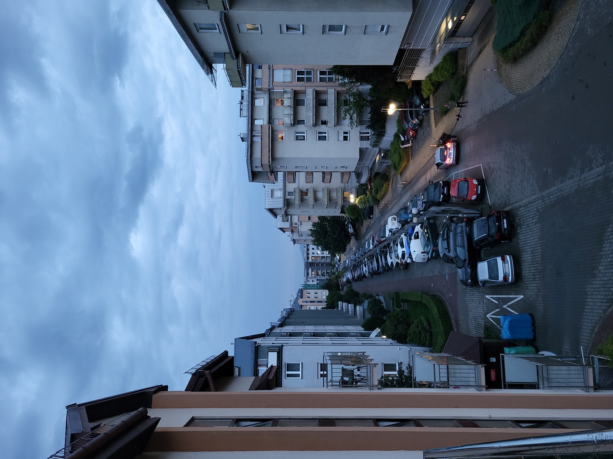 The image captures an urban residential scene at dusk or early night, characterized by an overcast sky filled with dark blue-grey clouds. The perspective is from an elevated position, likely from a building's window or balcony, looking down and across a street lined with apartment buildings.
In the foreground, the roofline and upper stories of a building with brown and beige facades are partially visible. Across the street, several multi-story residential buildings of varying modern architectural styles stand, some featuring balconies. Lights are illuminated in numerous windows, suggesting occupancy.
The street below is paved and features designated parking areas. A prominent scene in the midground involves a silver car with its brake lights illuminated and the driver's side door open. Two individuals are visible near this car; one person appears to be standing beside the open car door, while the other stands slightly further back on the pavement. The street is lined with numerous parked cars, extending into the background. Streetlights are illuminated, casting light on the pavement and surrounding areas.
Green landscaping, including bushes and trees, is integrated throughout the scene, bordering buildings and the street. On the ground near the street, two distinct, possibly tarp-covered objects, one blue and one green, are visible. A white 'X' marking is painted on the asphalt. The overall impression is that of a quiet evening in a densely populated urban or suburban environment. No discernible text is visible.