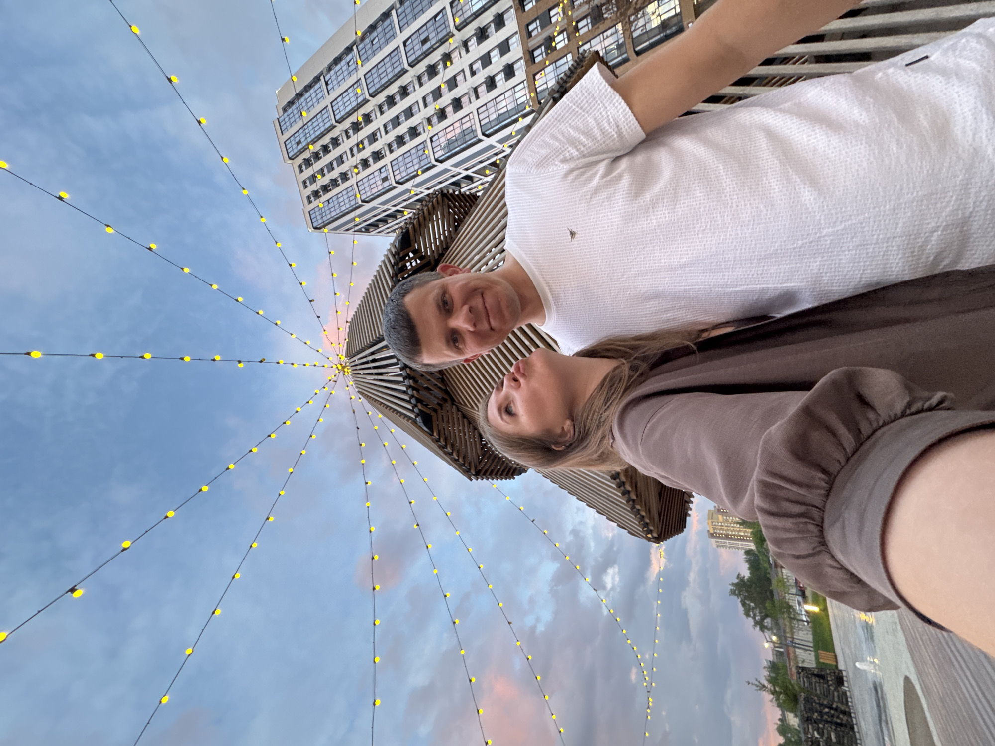 A man and a woman pose for a selfie against the backdrop of the city of Perm, Russia. The camera angle is from below, looking upward. The man wears a white, short-sleeved shirt. The woman is in a brown, long-sleeved garment. Behind them, a wooden tower structure with radiating string lights extends into a cloudy sky. A tall building with many windows rises to the right. In the distance, a park-like area with trees and other buildings is visible.