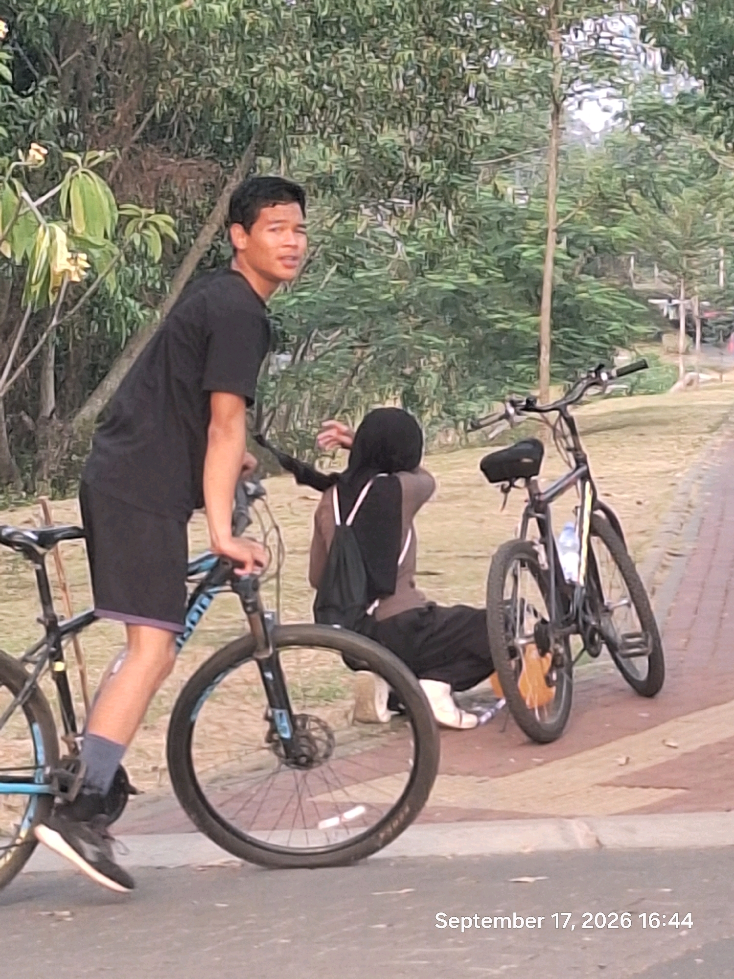 The image depicts an outdoor scene, likely in a park or recreational area in Depok, Indonesia. A young man, wearing a black t-shirt and black shorts with a purple trim, stands next to a bicycle, holding its handlebars. He is looking to his right. Behind him, a person wearing a black hijab and a brown top, with black pants, is seated on the ground next to another bicycle. The bicycles are dark-colored with some blue accents on the front bicycle. The environment features trees with green foliage and some lighter-colored flowers, a paved pathway, and a grassy area. The date and time stamp "September 17, 2026 16:44" is visible at the bottom right of the image.