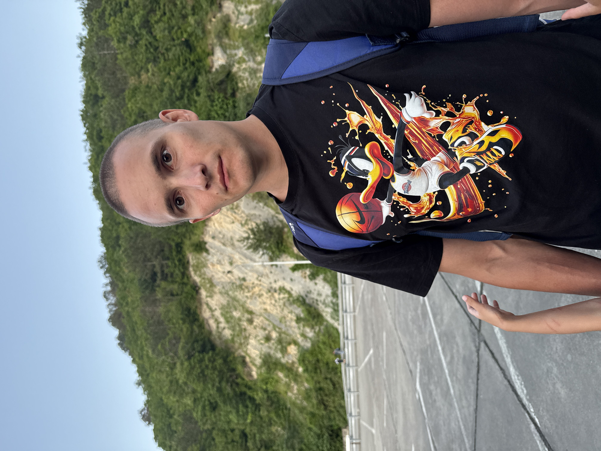 A man with a buzz cut and a black t-shirt featuring Daffy Duck playing basketball stands in a parking lot. A blue backpack is visible over his shoulders. A child's arm is reaching into the frame from the left. The background consists of a wooded hillside and a bright blue sky. The location is identified as Nebug, Russia.