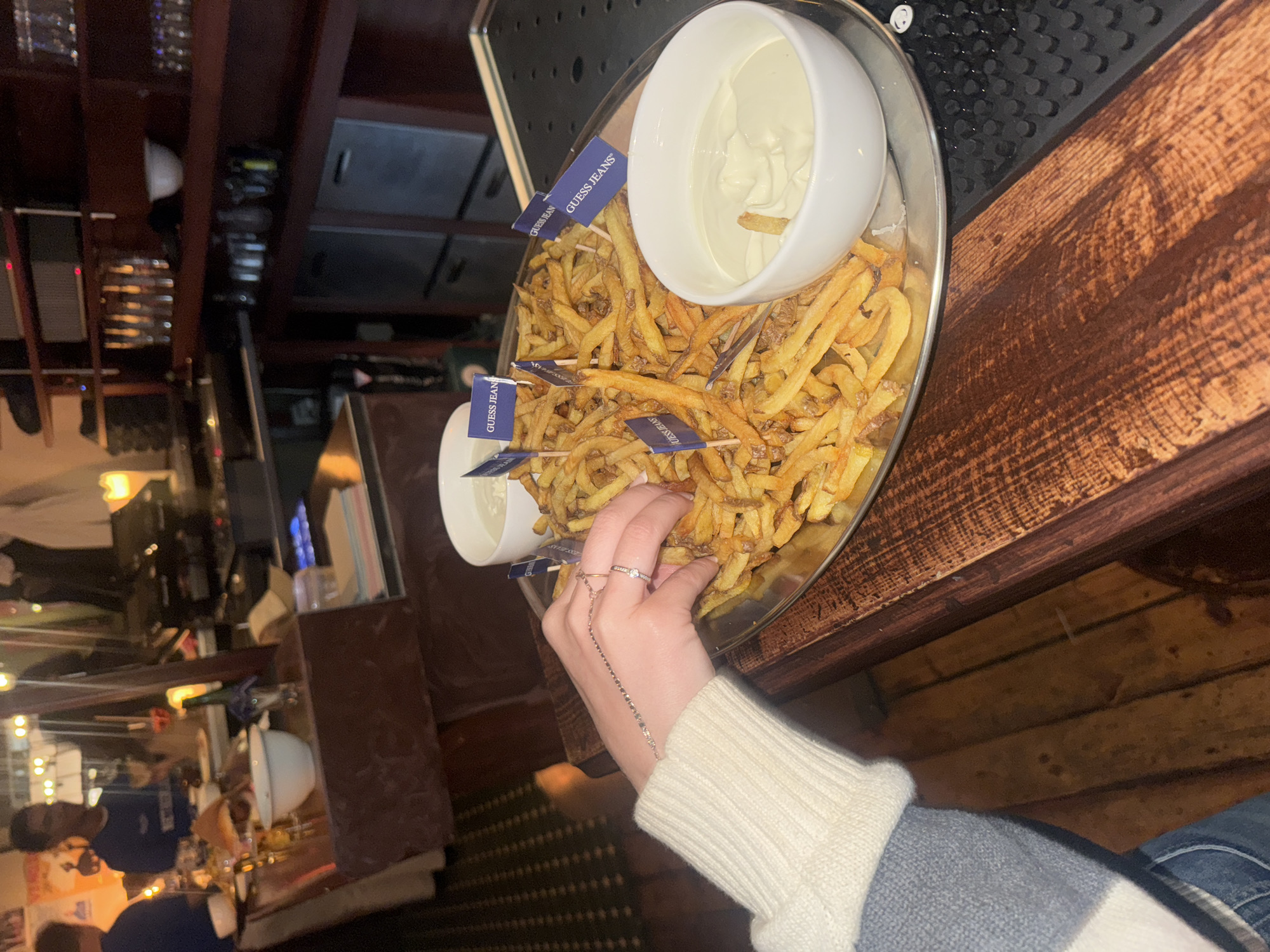 A woman's left hand, adorned with rings and a bracelet, reaches into a large platter of french fries at a dimly lit bar. The fries are scattered and some are adorned with small blue flags that say "GUESS JEANS". Two white bowls filled with a white dip are visible, one on the platter and one slightly behind it. In the background, the bar area is visible with shelves stocked with glasses and bottles, as well as some people in the distance. The overall setting suggests a casual bar or restaurant environment. The specific location in Amsterdam, Netherlands, cannot be determined from the visual information provided.