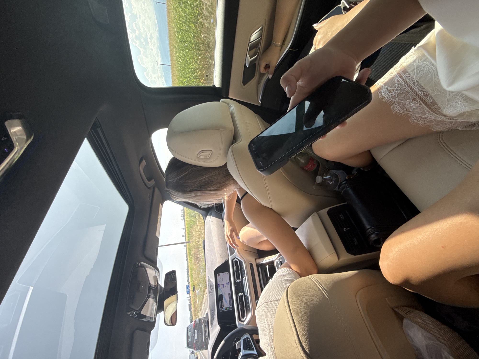 The image depicts the interior of a car, likely a modern sedan with tan leather seats and a sunroof, as seen from the rear passenger seats. Two women are present. The woman in the foreground is holding a smartphone, her legs extended into the frame. She is wearing a white garment with lace trim. The second woman is seated in the front passenger seat, facing away from the camera, with her arm resting on the center console. The car is driving on a highway, indicated by the road markings and other vehicles visible through the windows. The landscape outside consists of a field of crops, likely corn, under a bright sky with scattered clouds. Power lines are visible in the distance. The car's dashboard with a navigation screen is also partially visible. Given the context of the road, fields, and a cloudy sky, the location is Arpașu de Jos, Romania.