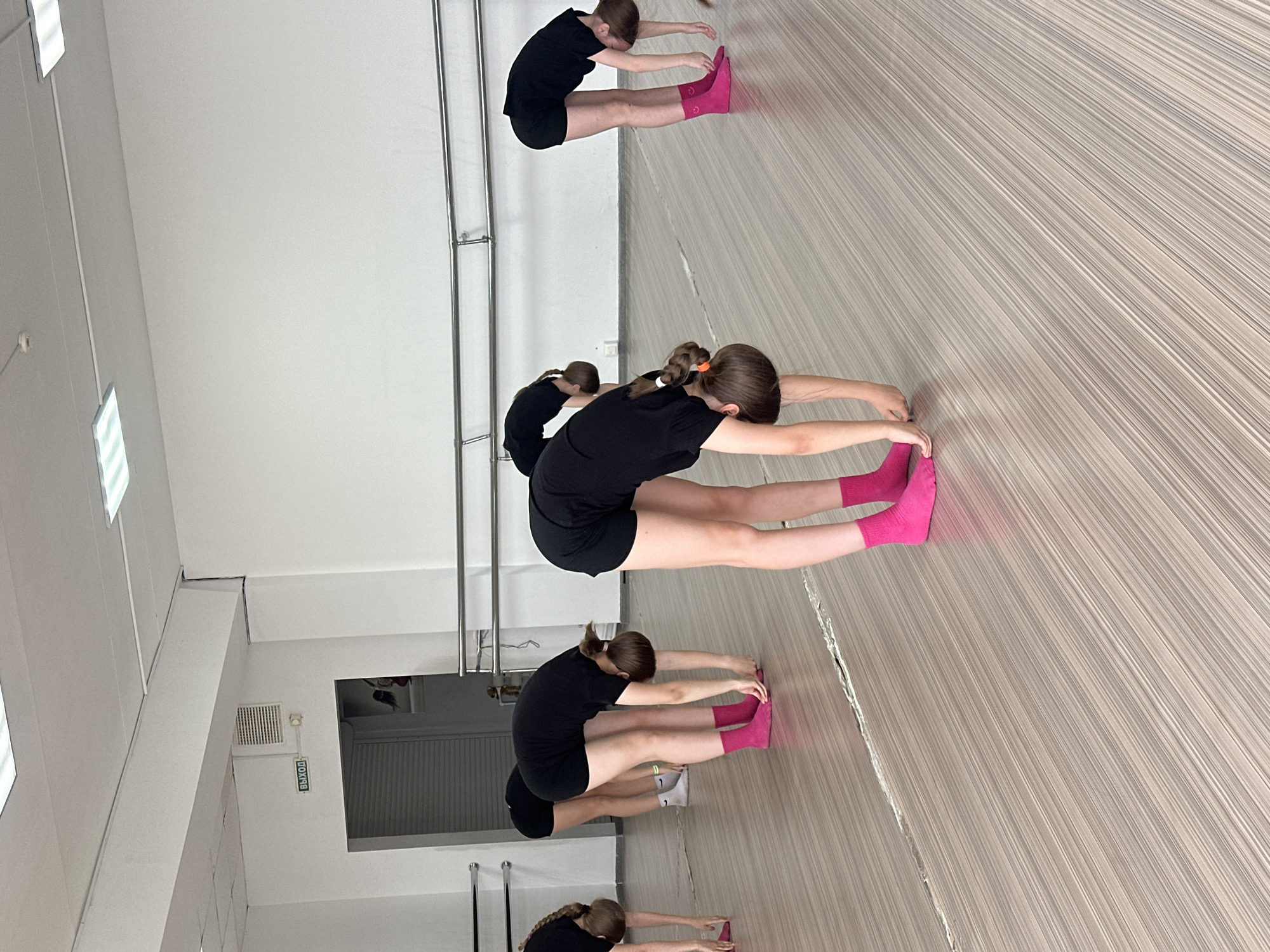 In an indoor dance studio in Votkinsk, Russia, four young girls are practicing stretching exercises. They are wearing black t-shirts and shorts with pink socks. They are seated on a light brown striped floor, stretching forward, and touching their toes. A silver ballet barre is along the wall behind them. The studio has white walls and a bright interior. An exit sign is also visible.