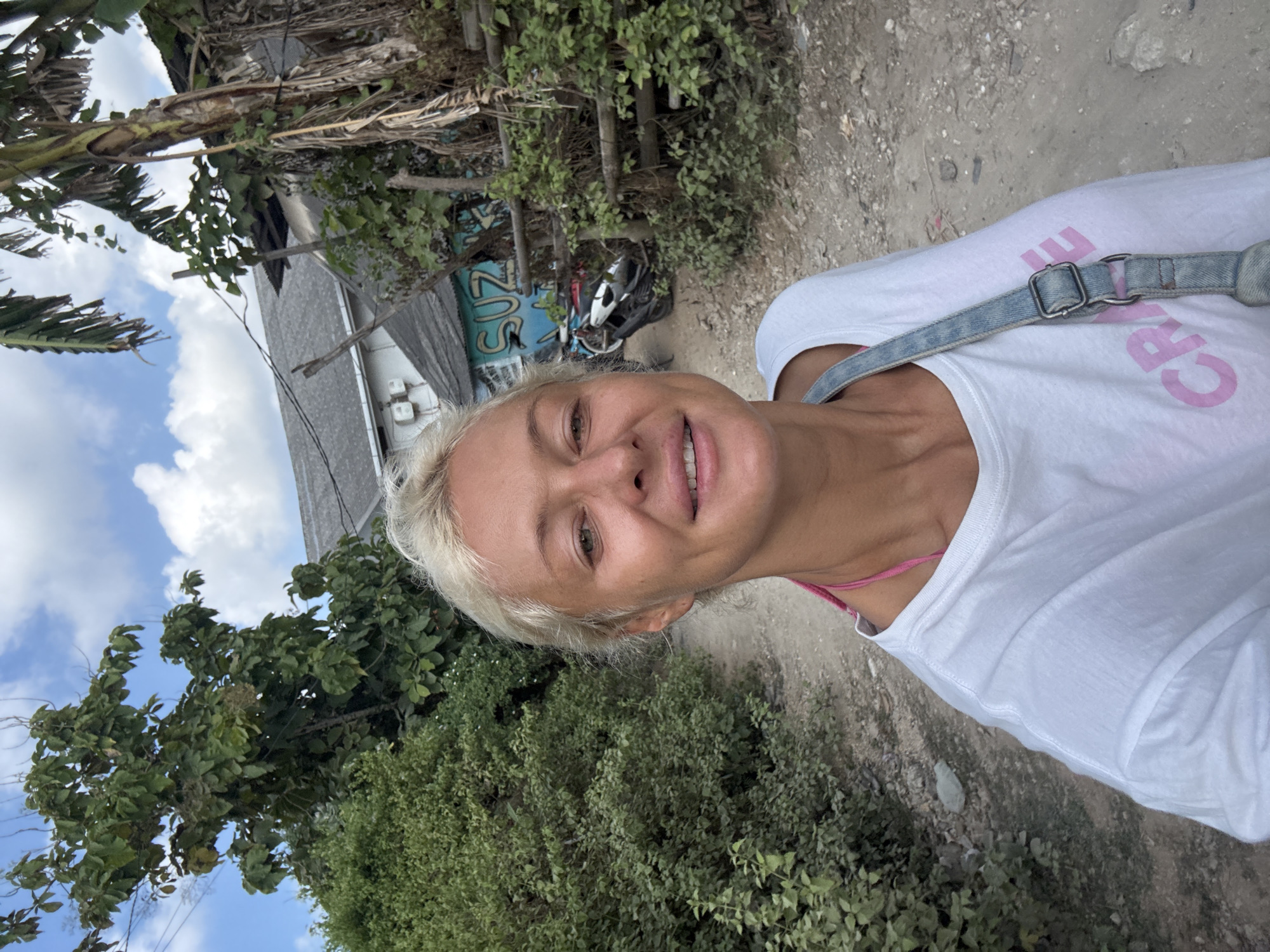 A blonde woman is taking a selfie outdoors. She is wearing a white t-shirt with pink text on the front and a denim strap across her chest. She has on a pink strap underneath her shirt. The background shows lush greenery, a dirt path, and some buildings with a motorcycle parked in front of one. The sky is partly cloudy.