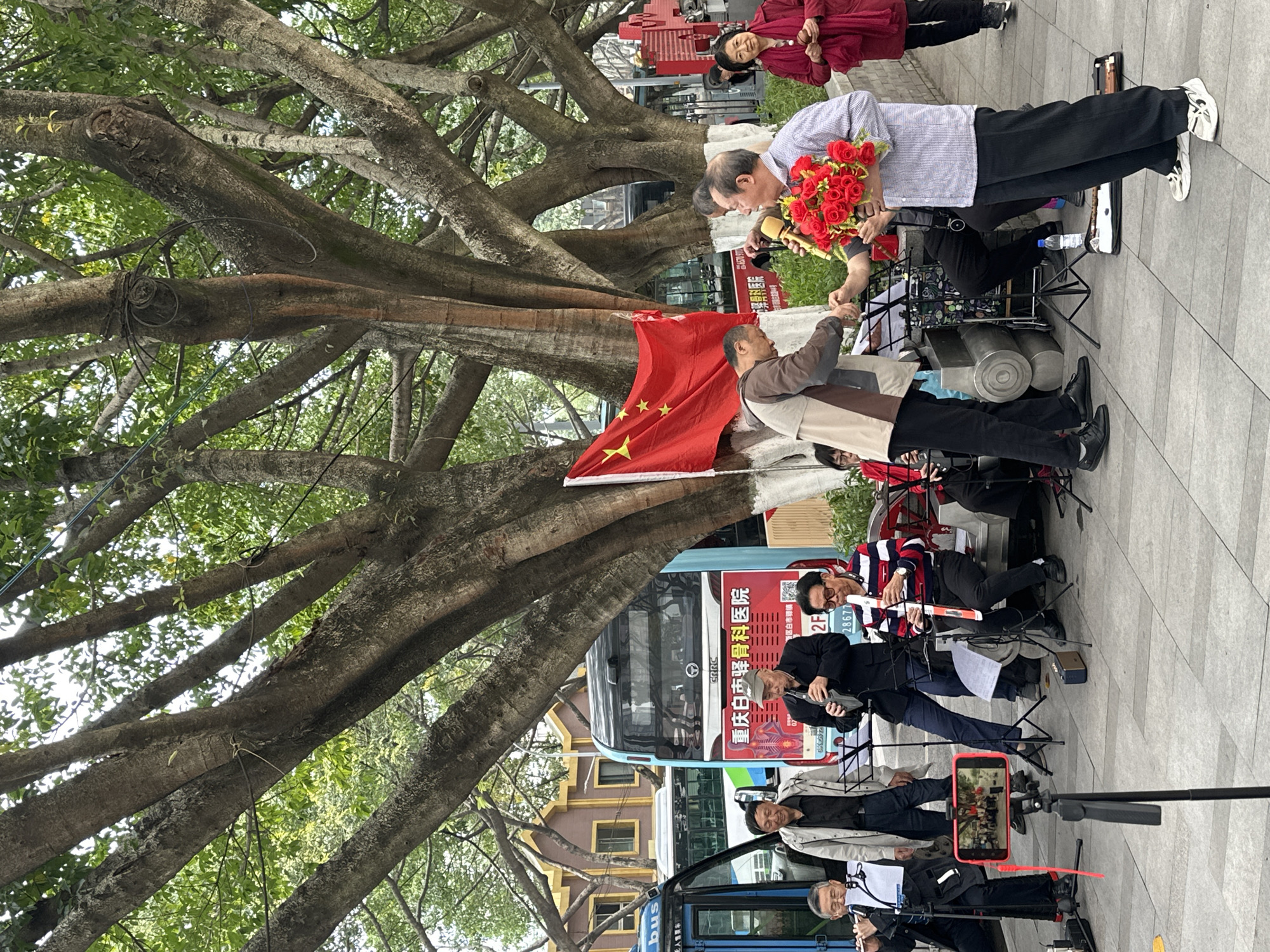 In the Jiulongpo District of China, a scene unfolds outdoors. A large tree dominates the foreground, its branches intertwined, displaying a Chinese flag. Two people stand facing each other, one holding a bouquet of red roses. Behind them, a small band is playing music. A bus with Chinese characters and the words "骨科医院" and "白市驿镇" is visible, along with several buildings in the background. A person in a red jacket stands to the side. The band's performance is being recorded on a smartphone.