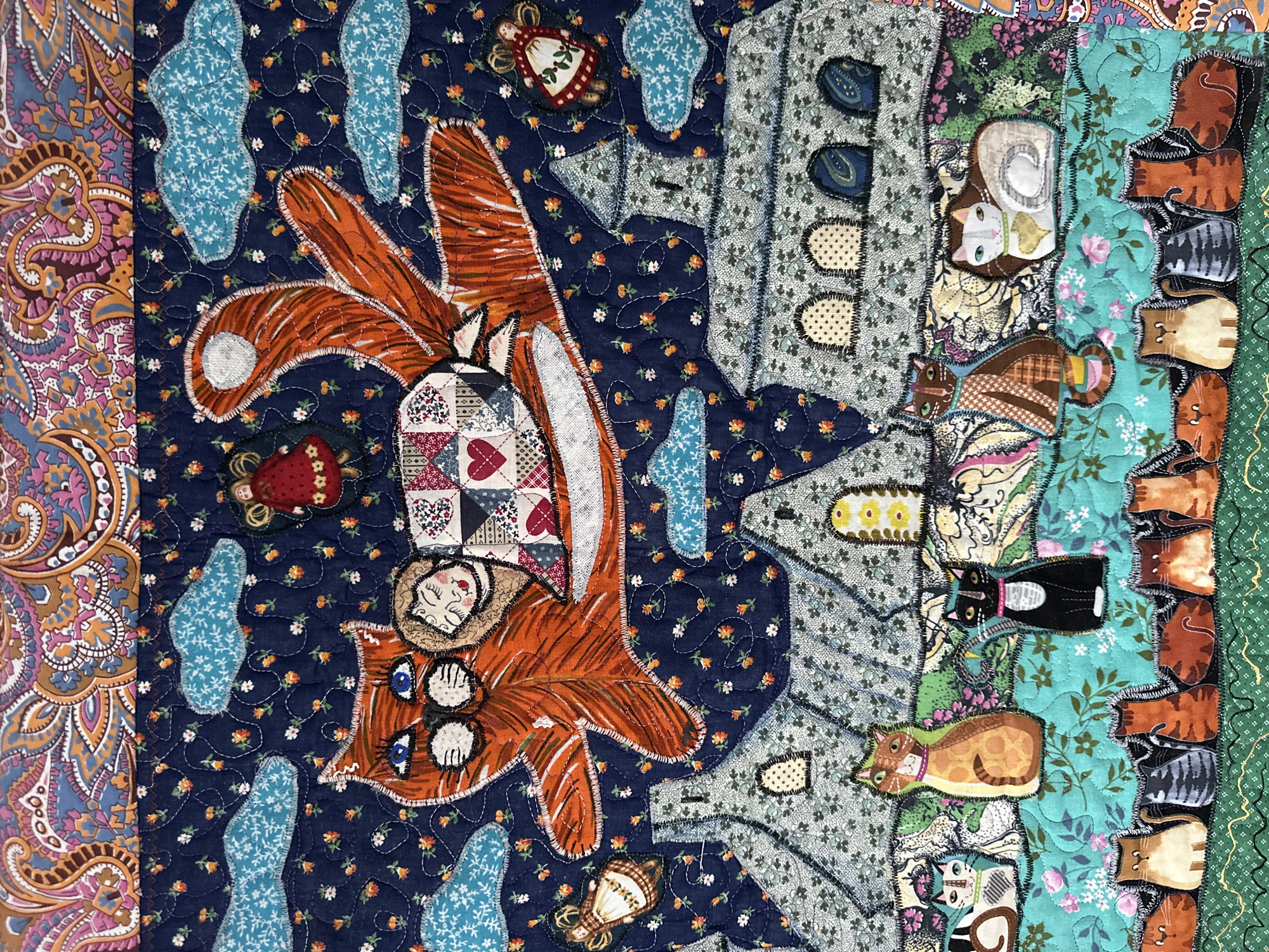 The image is a close-up, overhead shot of a colorful, handmade quilt. The quilt features a whimsical, dreamlike scene with a large orange cat flying through a dark blue, star-and-flower-patterned sky with light blue clouds. A small, blonde-haired doll rests on the cat's back, wrapped in a patchwork blanket with a red heart. Below the flying cat are stylized buildings that resemble churches or castles, made from patterned gray fabric. In the foreground, a row of fabric cats of various colors and patterns sits on a teal, floral-patterned background. Below this row, another line of smaller, simplified cat shapes are arranged horizontally. The top portion of the quilt displays a large, paisley-patterned fabric in shades of orange, pink, and blue. The quilt is intricately stitched, with visible thread work outlining the shapes. The scene depicted is entirely fantastical and does not contain any elements that would suggest a specific location in Moscow, Russia, or any real-world urban environment.