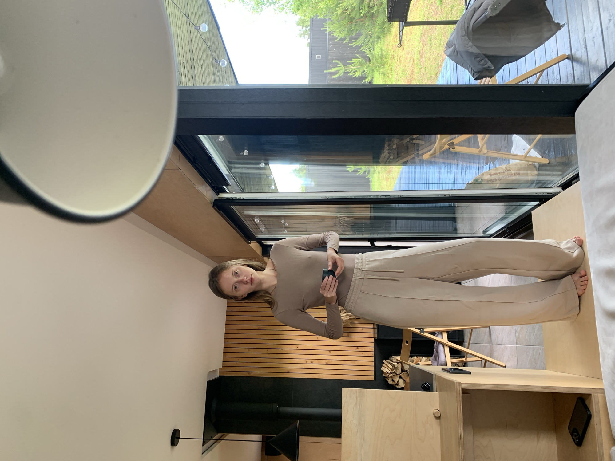 A young woman stands in a modern, minimalist interior with large glass doors looking out onto a backyard. She is wearing a fitted, long-sleeved beige top and loose-fitting beige pants. She holds a small, dark teal object in her hands. The room features natural wood accents on the walls and cabinetry, a black stovepipe, and a black pendant light. Through the glass doors, a wooden deck, green grass, trees, and a dark fence are visible. A chair and a grill can be seen on the deck.