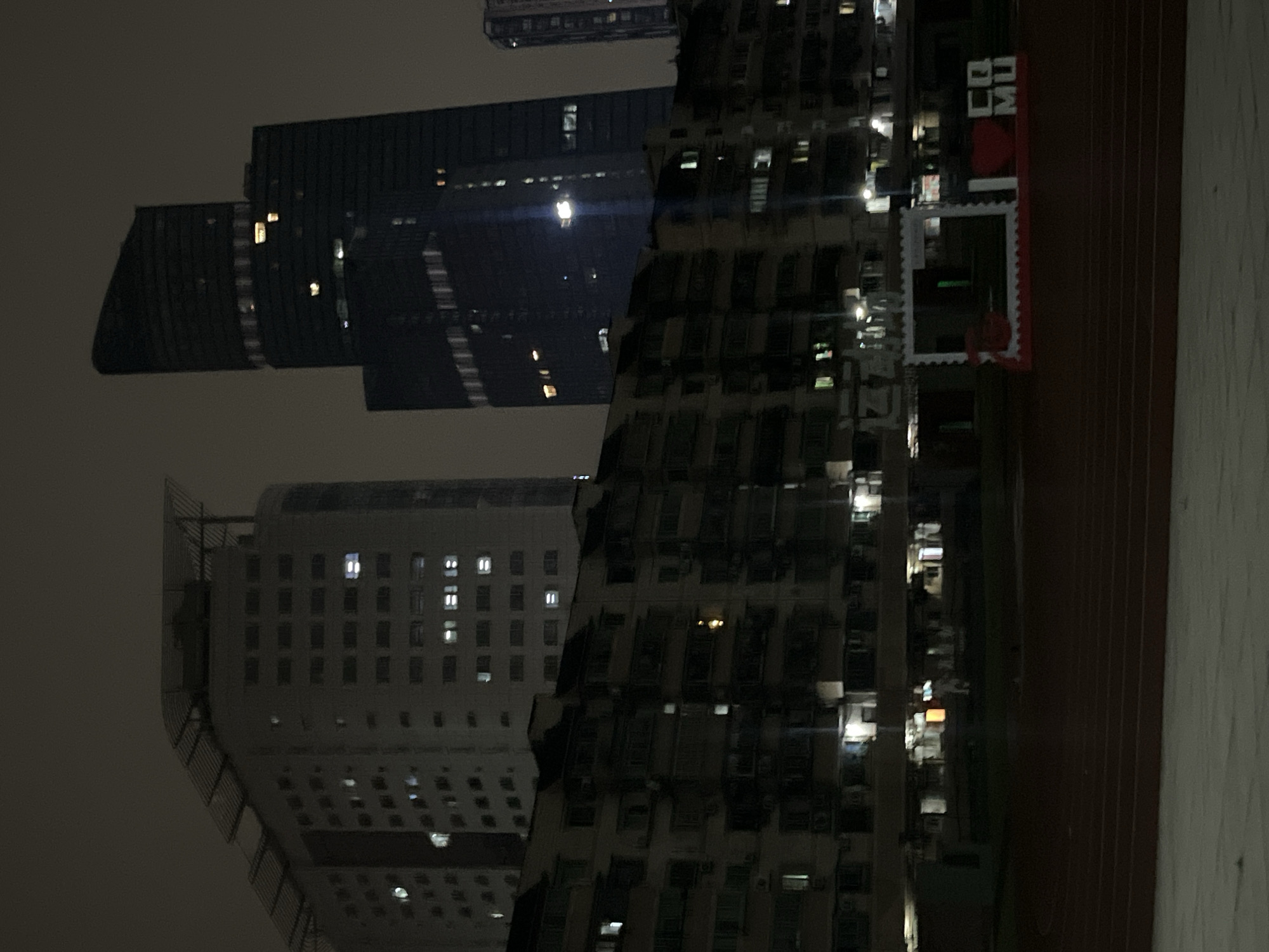The image depicts a nighttime scene in Yuzhong District, China, featuring a cluster of buildings and a track. In the foreground, a reddish-brown track is visible, with a white, illuminated structure in the middle distance. This structure is rectangular with decorative white edging and appears to be a photo frame or a stage decoration. Inside the frame, a red heart shape and white block letters spelling "CO MU" are prominently displayed.
Behind the illuminated structure, a low-rise apartment building stretches across the mid-ground. It is several stories tall with numerous windows, many of which are illuminated, suggesting occupancy. The building has a somewhat aged appearance with visible balconies and air conditioning units.
In the background, taller modern buildings dominate the skyline. To the left, a light-colored, multi-story building with a distinctive curved top and rooftop structure is visible. To the right of this, two dark, sleek skyscrapers rise high into the night sky. The left skyscraper is taller and has a more angular, modern design, while the right one appears slightly shorter with a more rounded top. Further to the right, another tall, slender building is partially visible. The overall atmosphere is urban and nocturnal, with the city lights reflecting off the building facades and illuminating the scene. The sky is dark and overcast, contributing to the nighttime ambiance.