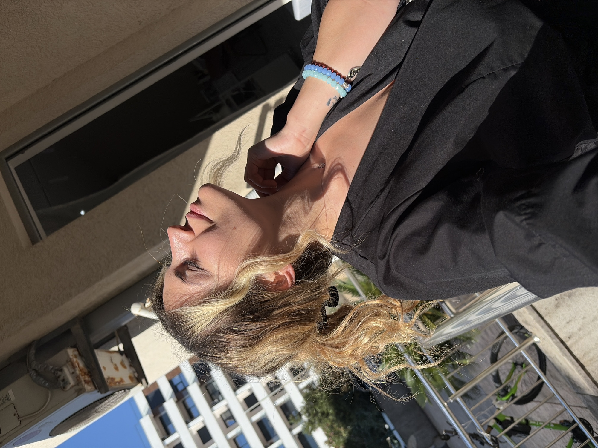 A woman with blonde and brown hair is posing outside on a balcony, in Budva, Montenegro. She wears a black blouse and two beaded bracelets on her left wrist. Her hand is raised near her neck. The balcony has a metal railing, and a bicycle is visible below. A multi-story building is in the background, along with an air conditioning unit.
