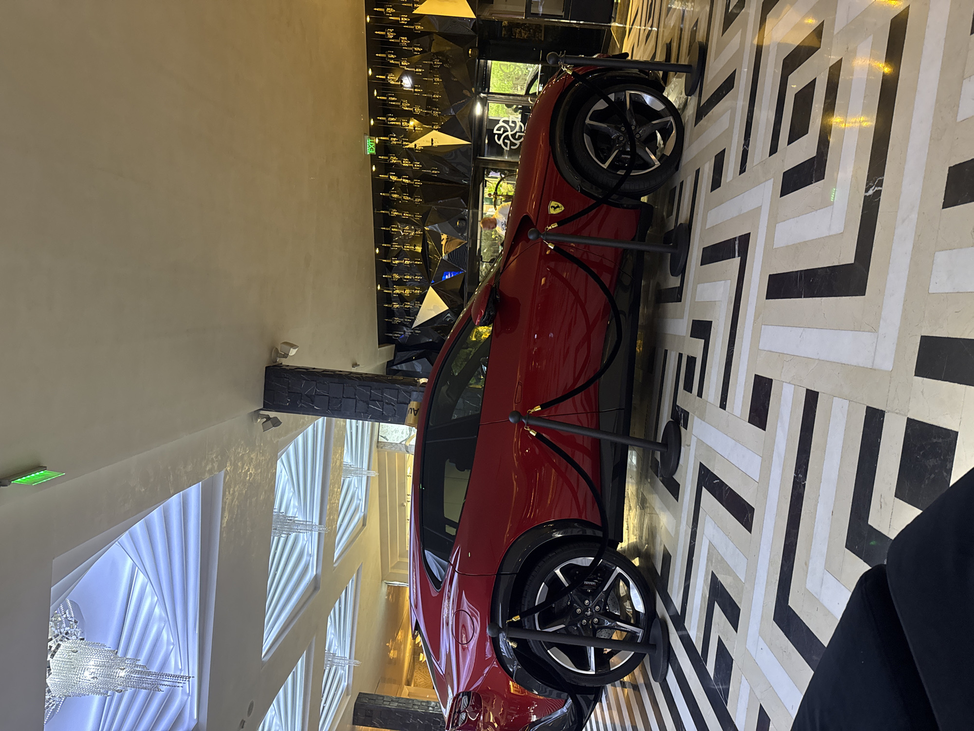 An indoor scene showcases a red luxury sports car, appearing to be a Ferrari Purosangue, prominently displayed on a geometrically patterned floor. The car is positioned on a black, white, and beige tiled floor with an interlocking angular design, and is roped off by black velvet ropes held by black stanchions, suggesting an exhibition or showroom setting.
The main subject is the sleek red vehicle, visible from its rear three-quarter view, with its distinctive Ferrari emblem faintly visible on the side and clearly on the wheel hub. The car features black multi-spoke wheels with large brake calipers.
The surrounding environment is characterized by luxurious and modern decor. The walls on the left are a plain, light color, while the back wall features intricate black cladding with a faceted, triangular design, adorned with numerous small, glittering golden-hued decorative elements that resemble bullet casings or small bottles. A green "EXIT" sign is visible on the far left wall. On the right side, the floor pattern extends, and a person's leg, clad in black, is partially visible in the bottom right corner, suggesting someone is seated or standing nearby, observing the display.
The ceiling area, visible towards the top left, features elaborate crystal chandeliers hanging in recessed, wave-like or spiraling light fixtures, contributing to the upscale ambiance. Reflections of these light fixtures and other elements are visible on the car's glossy surface. The lighting is bright and artificial, illuminating the car and the surrounding space effectively. The overall setting implies a high-end venue, possibly a hotel lobby or a luxury dealership in Sofia, Bulgaria.