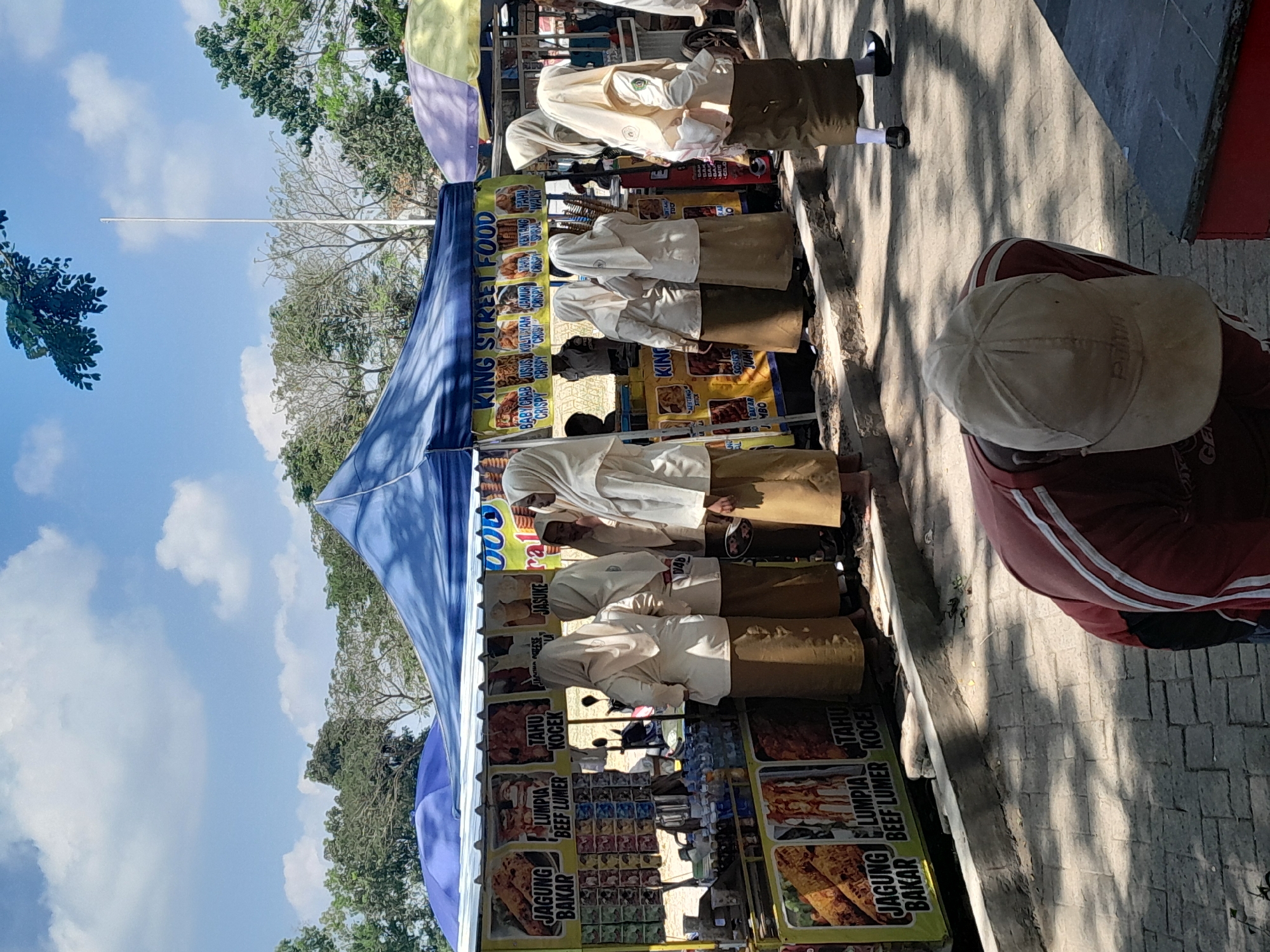 In Genteng Kulon, Indonesia, an outdoor food stall is set up under a blue tarp, displaying various food items with Indonesian text such as "JAGUNG BAKAR" (grilled corn) and "LUMPIA BEEF LUMER" (beef spring rolls). Several young females, dressed in light-colored uniforms and hijabs, stand near the stall, some appearing to interact with each other or examine the food offerings. Another person, wearing a red shirt and a light-colored cap, is partially visible in the foreground, bending over. The environment is sunny with a clear blue sky and scattered clouds, and trees are visible in the background. The ground is paved with brick-like material.