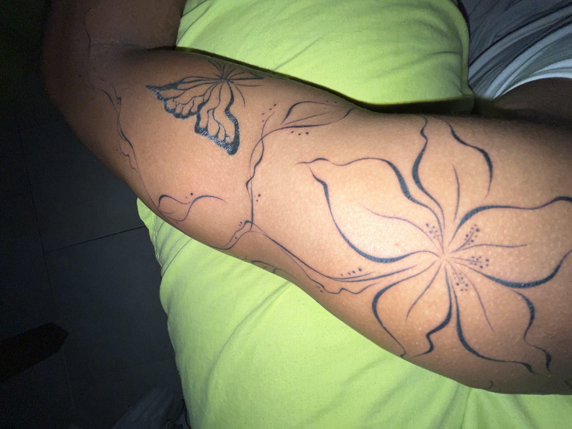 A close-up shot of a person's arm with black line tattoos of a flower and a butterfly is featured against a bright green fabric. The tattoos are in various stages of completion, with some lines bolder than others. The person's skin tone is dark. The background is dark and out of focus, with a hint of striped fabric at the bottom. The image was taken in La Asunción, Venezuela.