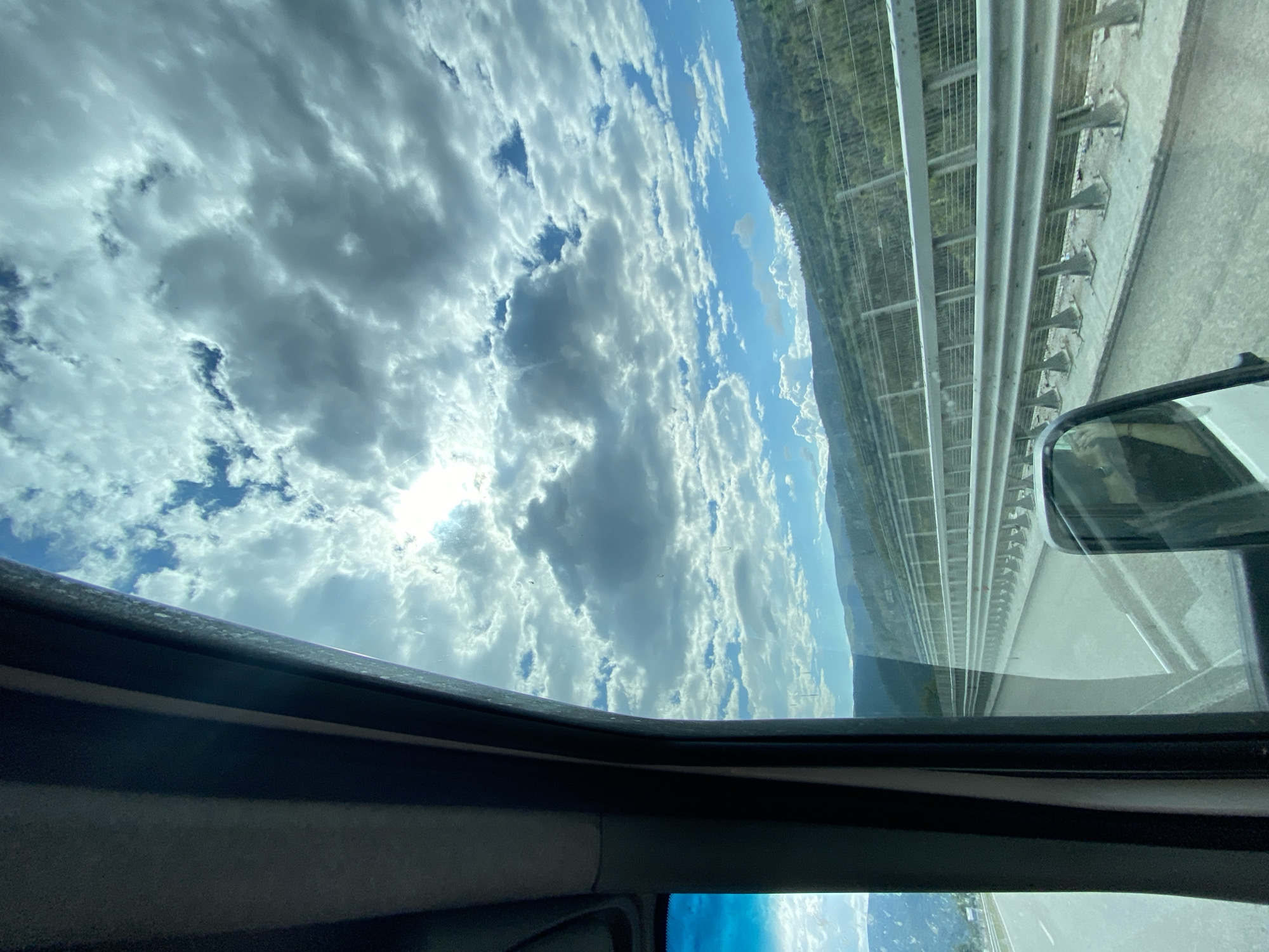 The image, taken from inside a vehicle, captures a view through the windshield and side window. The foreground is dominated by the interior of the vehicle, with a dark-colored pillar and a portion of the dashboard visible on the left. The right side of the image shows a side mirror reflecting a blurred image of a person.
The external view is a landscape with a highway stretching into the distance. A metal guardrail lines the right side of the highway. Beyond the highway, a densely forested hillside rises, suggesting a rural or mountainous setting. The sky is filled with a dramatic display of clouds, with the sun partially visible behind them, casting bright light. The overall impression is of a journey through a scenic route, likely a highway in or near Vrachesh, Bulgaria, given the context. The time of day appears to be daytime, possibly late morning or early afternoon, due to the bright sunlight.