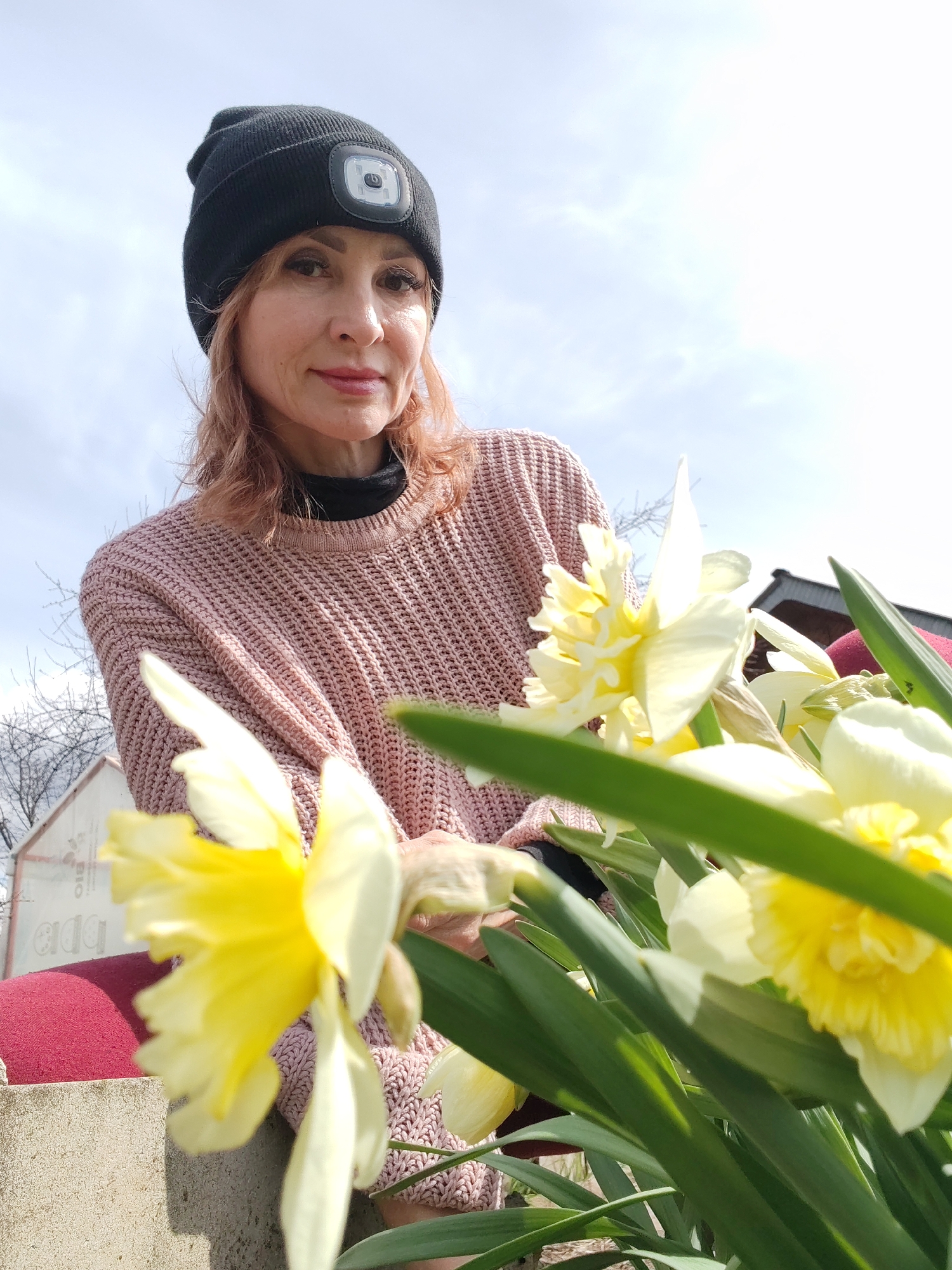 A woman is pictured in an outdoor setting in what appears to be a backyard or garden, likely in the city of Chaikovsky, Russia. She is wearing a black beanie with a light attached, a pink knitted sweater over a black turtleneck, and is positioned behind a cluster of yellow and white daffodils. The background includes a glimpse of a building, possibly a shed or house, and a sky with light clouds. The woman is looking toward the camera. The lighting is bright, suggesting it is daytime. A red cushion is visible under the flowers.