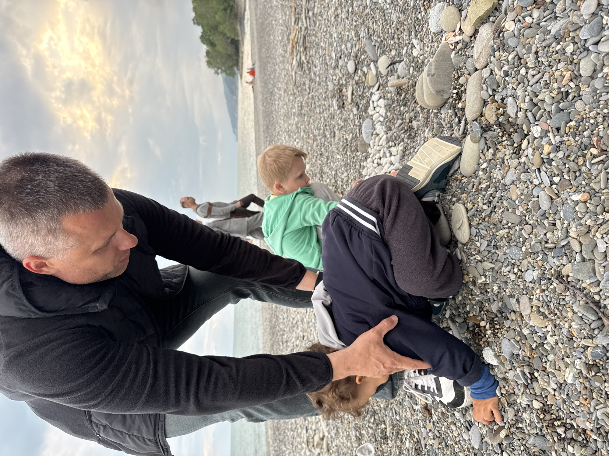 On a pebble beach in Sochi, Russia, a man in a black jacket and gray pants is adjusting the position of a child wearing a dark blue jacket, gray hoodie, and black and white shoes. The child is bent over, seemingly looking at the pebbles. Another child in a light green shirt sits nearby. Two other adults are lying on the beach in the background. The sky is cloudy, and trees are visible in the distance.