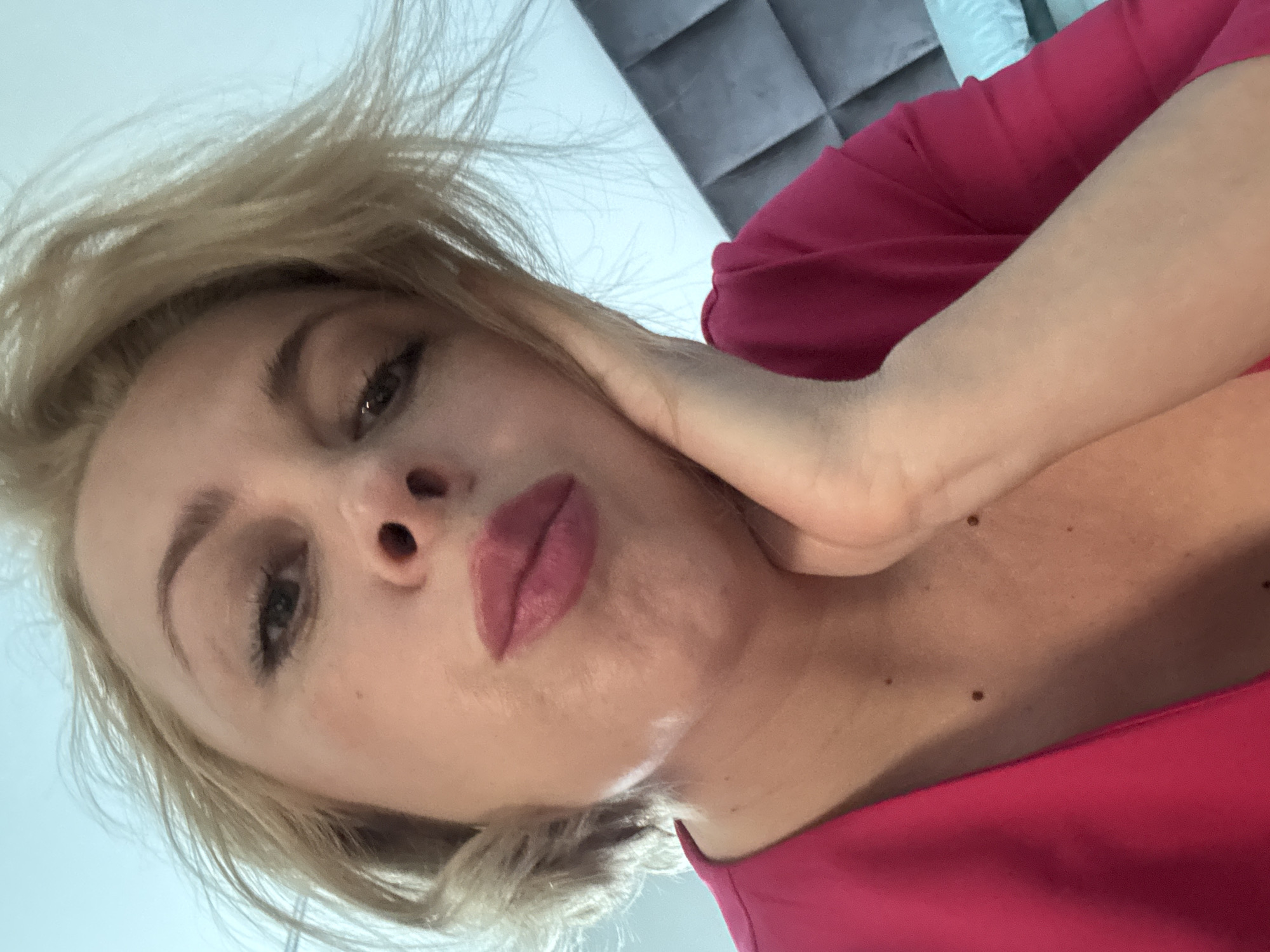 A woman with blonde hair and pink lipstick is looking down at the camera, with her right hand on her cheek. She is wearing a bright pink top. The background is out of focus, but appears to be a room with a grey quilted headboard and a light blue pillow. The image does not contain information that places it in Sochi, Russia.