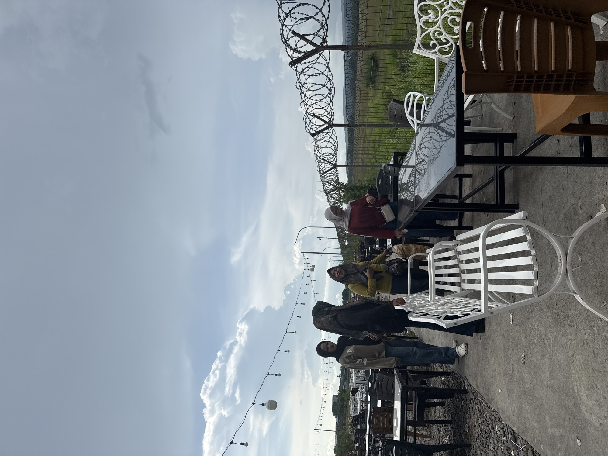 In Cotmalem, Indonesia, an outdoor cafe seating area is captured under a cloudy sky. Four women stand near a row of tables and chairs. One woman in a tan sweater and black hijab, and another in a black sweater and patterned hijab, are positioned to the left. A third woman in a yellow shirt and patterned hijab is in the center, holding a bag. The fourth woman, wearing a red shirt and light grey hijab, stands to the right. String lights with round bulbs are suspended overhead. A barbed wire fence runs horizontally across the background, separating the cafe from a grassy field and distant hills. The foreground features a concrete path with various outdoor furniture, including white metal benches, black tables with reflective surfaces, and an orange plastic chair. The ground is covered in gravel and scattered debris.