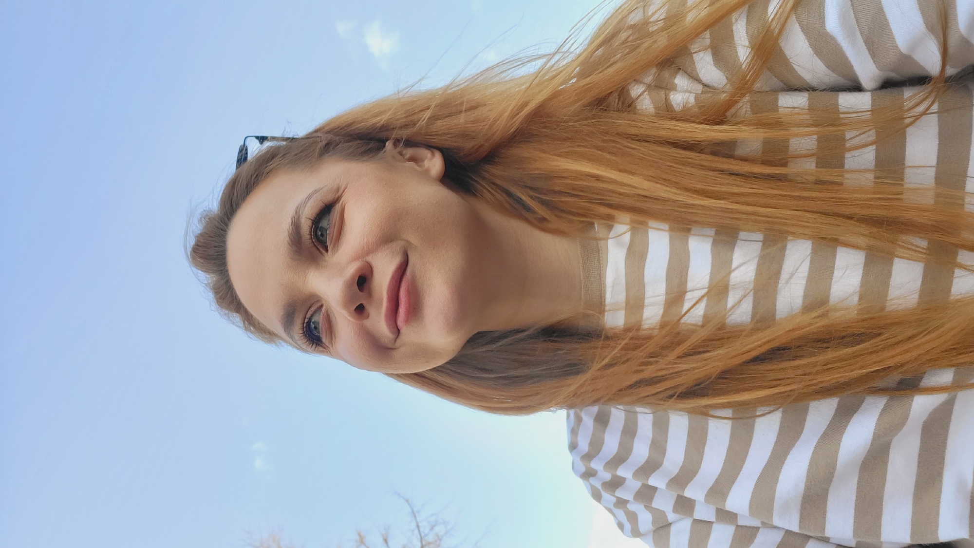 A woman with reddish-blonde hair, wearing a striped beige and white sweater, is positioned in front of a light blue sky. The image is taken from a low angle, looking up at the woman. The location is outdoors, possibly in the city of Chaikovsky, Russia, though specific landmarks are not visible to confirm this. The woman's expression is slightly smiling, and she appears to be looking slightly off-camera.