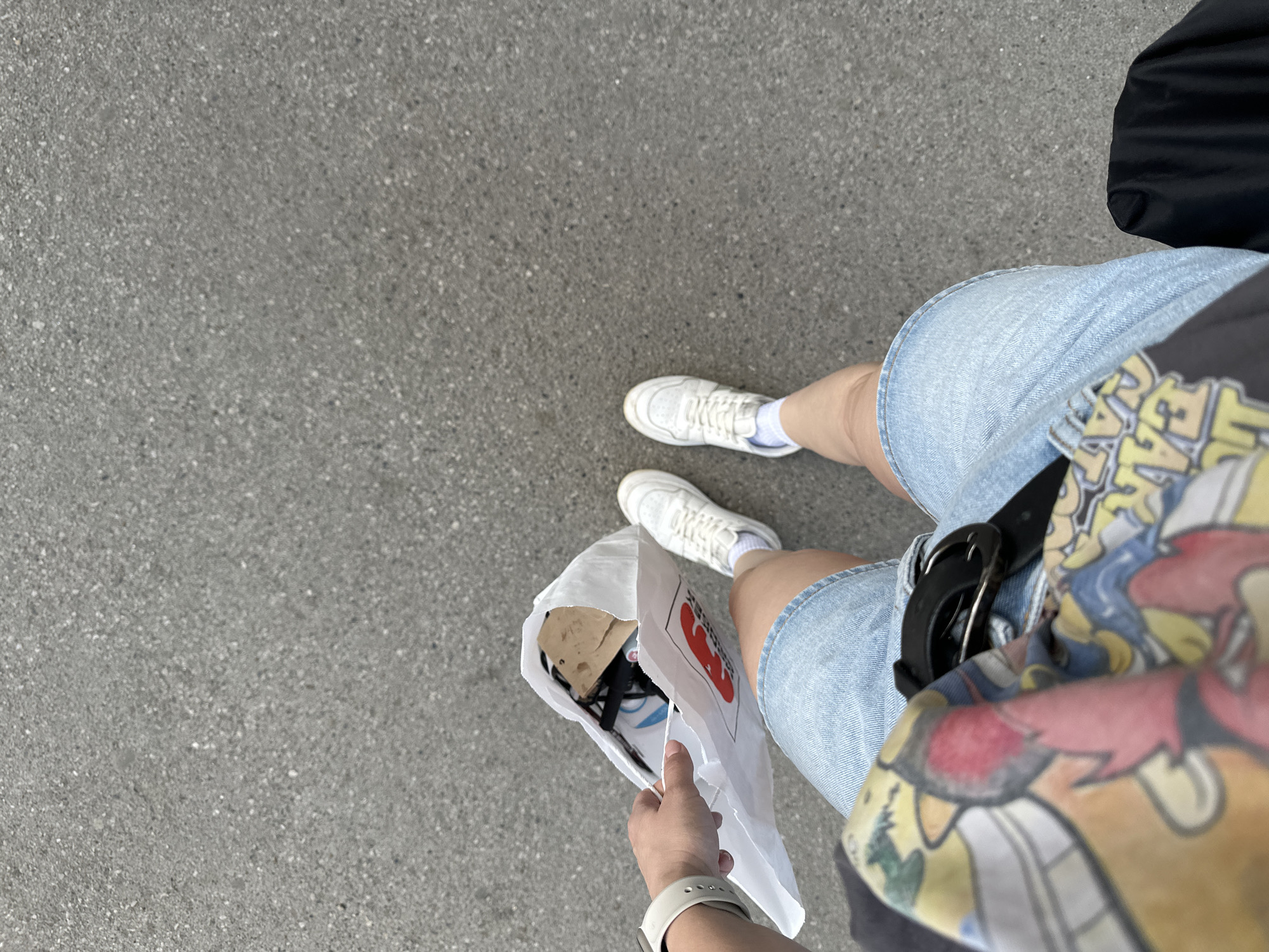 A high-angle medium shot depicts a person standing on a grey asphalt surface, holding a white paper bag with an indiscernible object inside. The person is wearing light blue denim shorts, a graphic t-shirt, and white sneakers with white socks. The person's left wrist is visible, adorned with a white smartwatch. A black bag is visible in the upper right corner of the frame. The image was taken in the city of Perm, Russia.