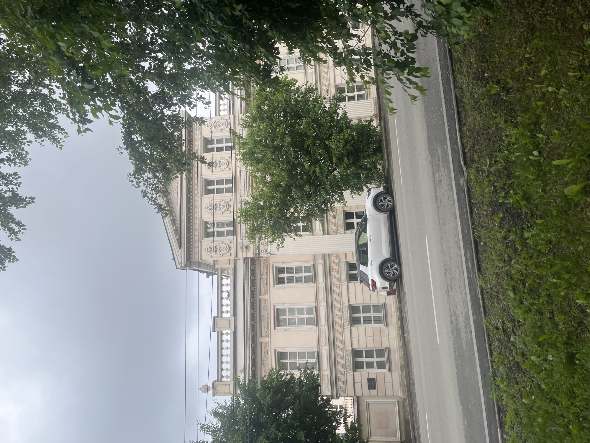 A white SUV is parked on the side of a street in front of a pale pink building. The building has a classical facade with columns, arched windows, and decorative carvings. Trees are present on both sides of the building and in the foreground, obscuring parts of the scene. The sky is overcast. The street is paved and has white lane markings. A strip of green grass and vegetation is visible along the edge of the road in the foreground.