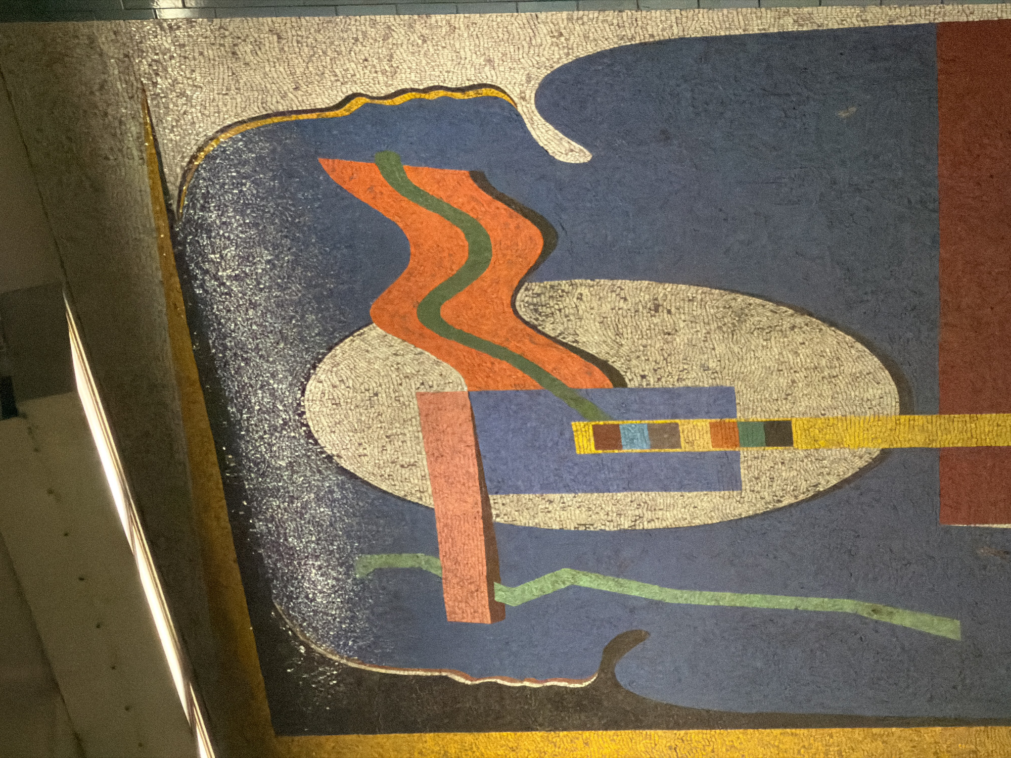 The image shows a colorful mosaic mural. It features geometric shapes in hues of orange, green, blue, white, yellow, and red. The shapes include a curved orange line with a green stripe running through it, set against a blue background, a white oval shape, a yellow horizontal rectangle, and a standing red rectangle. The mosaic has a textured appearance, and some sections are outlined in yellow and black. It is located in Atlanta, Georgia, United States of America.