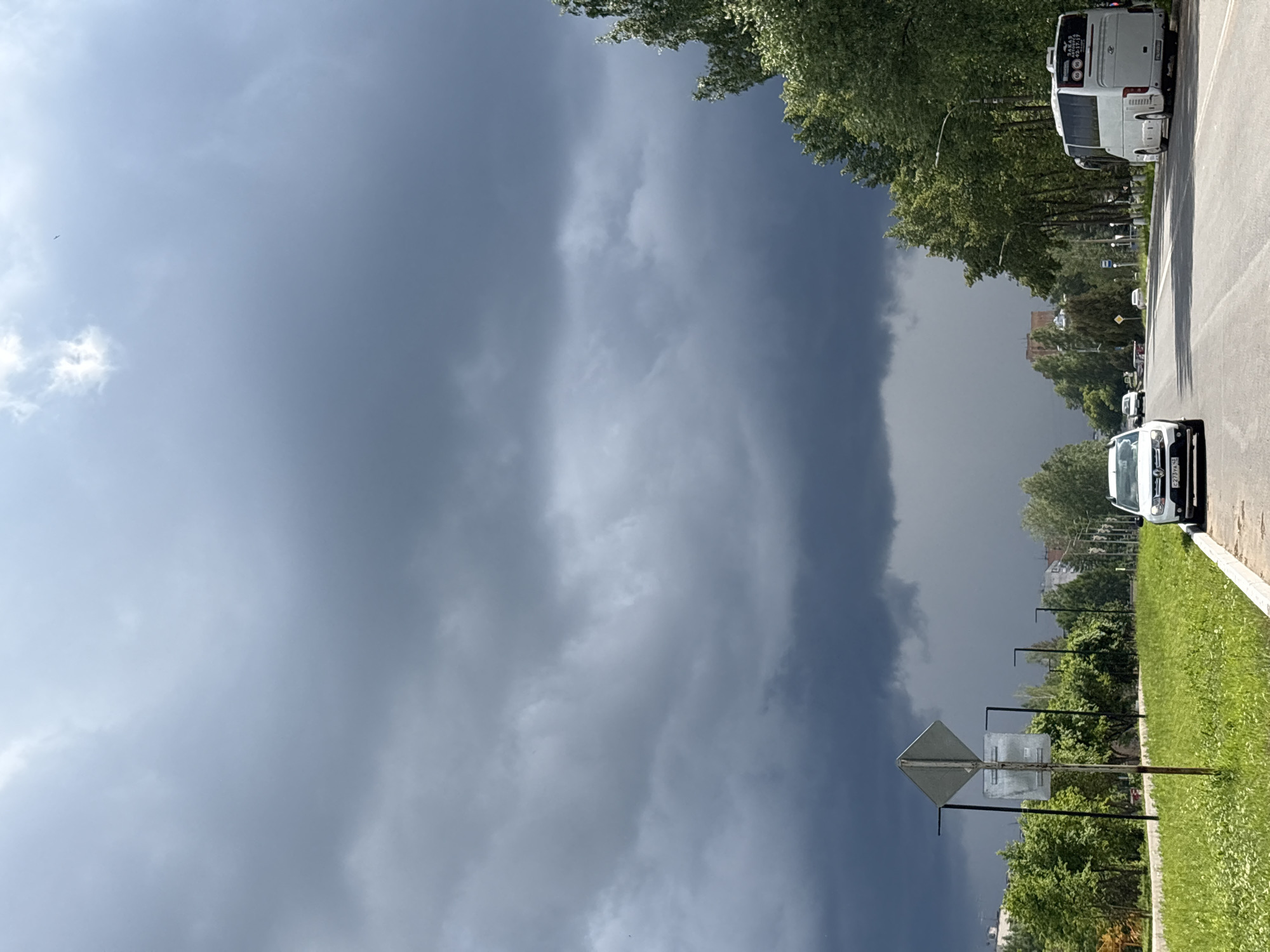 A wide shot shows a road in Kirovo-Chepetsk, Russia, under a dramatic, dark sky filled with storm clouds. On the right side of the road, a white bus is parked, and further down the road, a white car is traveling. The left side of the road has a grassy verge with trees and a traffic sign. Buildings can be seen in the distance.