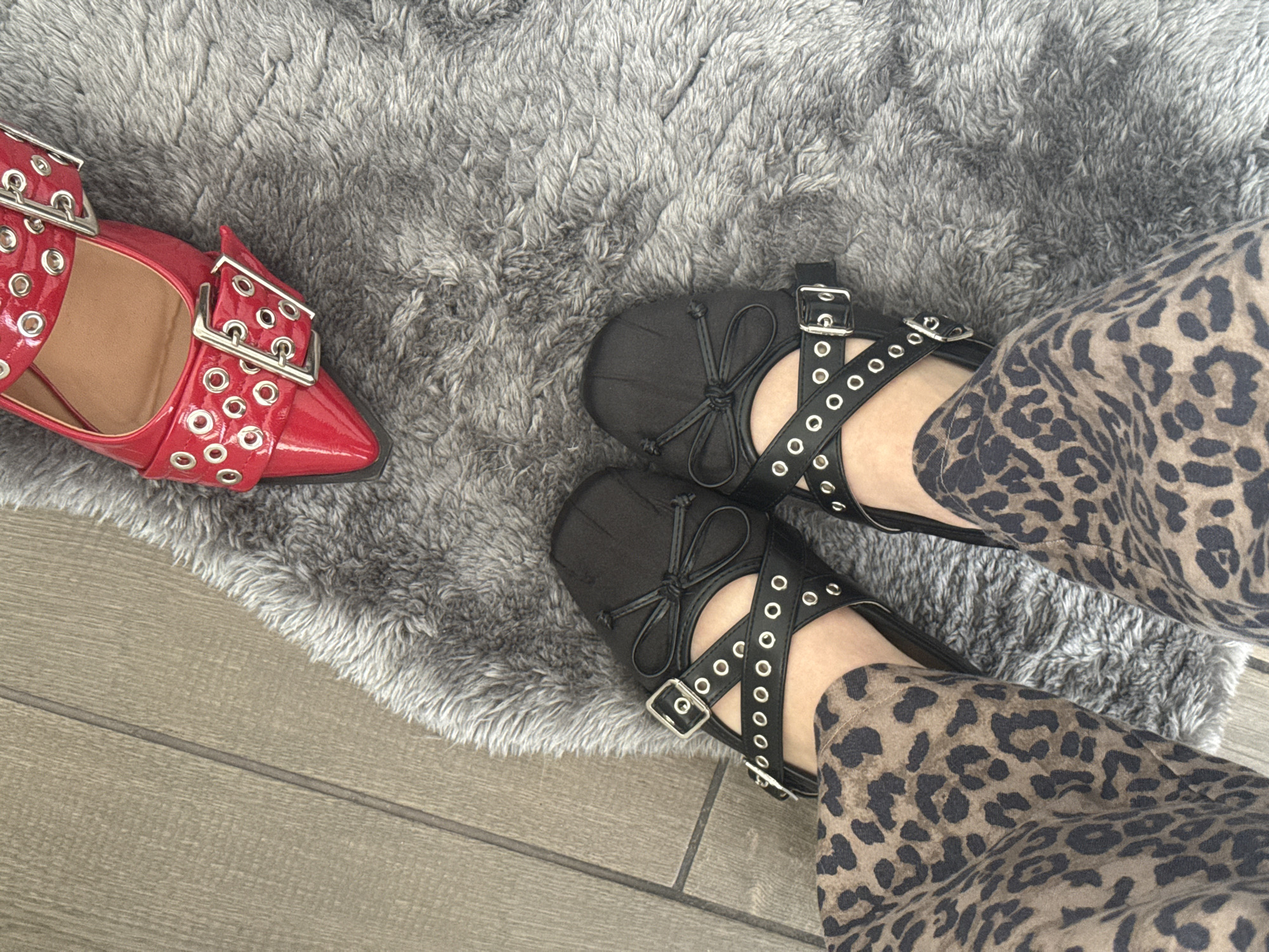 The image presents a close-up, overhead shot of a person standing on a gray, fluffy rug, with part of a wooden floor visible. The person is wearing leopard-print pants. On their feet are black flat shoes with decorative straps, buckles, and eyelets. Adjacent to the person’s feet, on the same rug, is a single red high-heeled shoe with similar buckle and eyelet details. The overall scene appears to be indoors, possibly a residence. There is no information to determine whether the photo was taken in Mexico City, Mexico.
